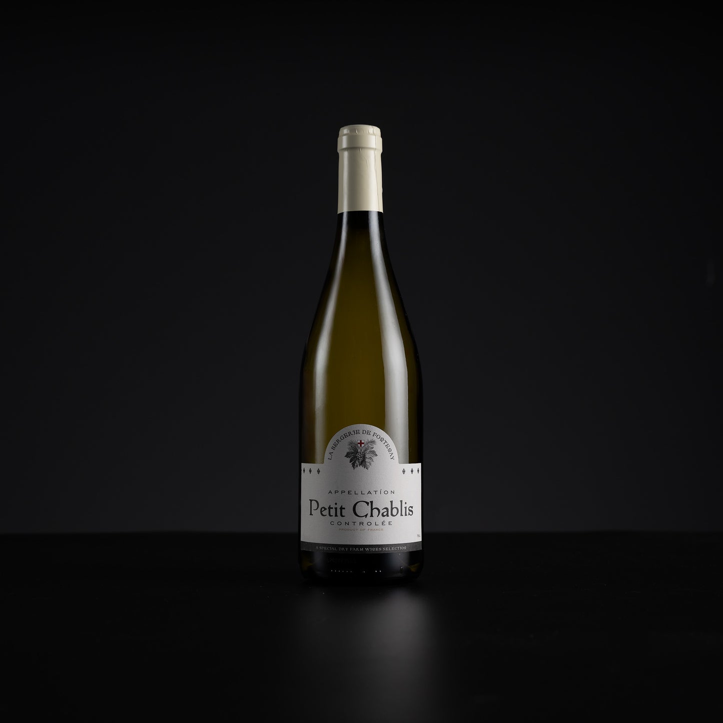 A single bottle of Petit Chablis white wine stands centered against a very dark black background, with a white label and pale foil at the neck.