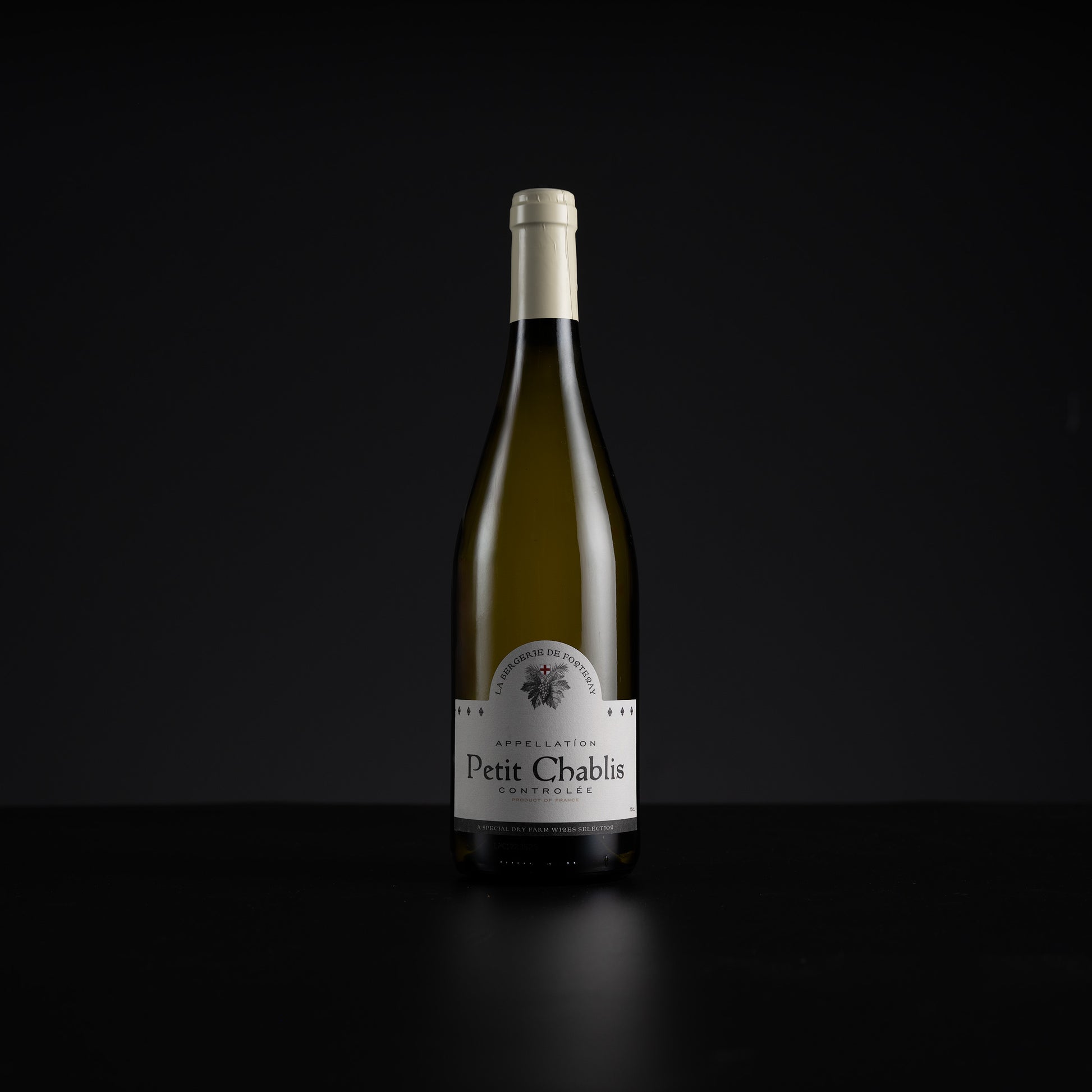 A single bottle of Petit Chablis white wine stands centered against a very dark black background, with a white label and pale foil at the neck.