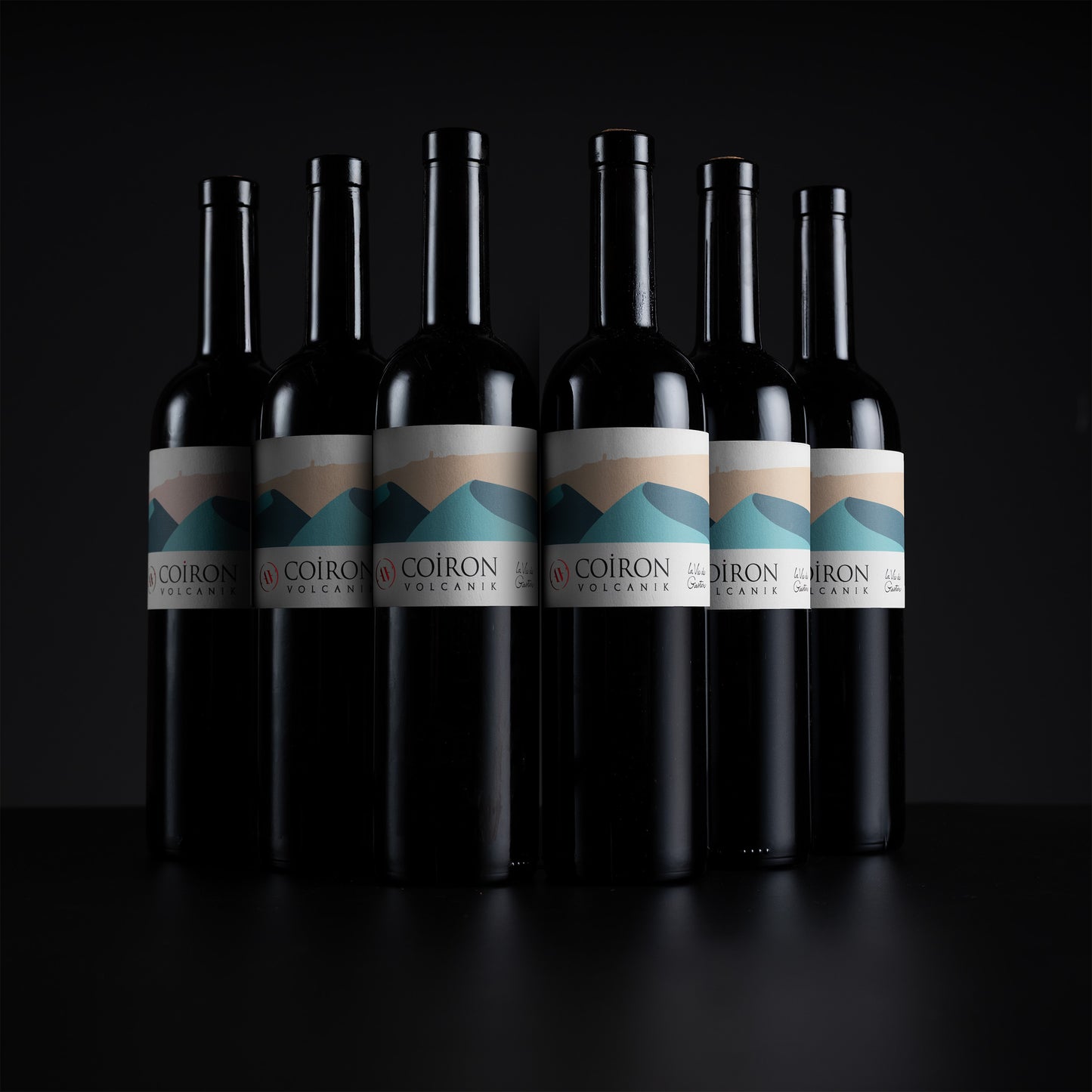 Six dark wine bottles with white labels featuring a blue and beige mountain graphic, arranged in a row against a black background. The label text reads 'COIRON OREGON'.