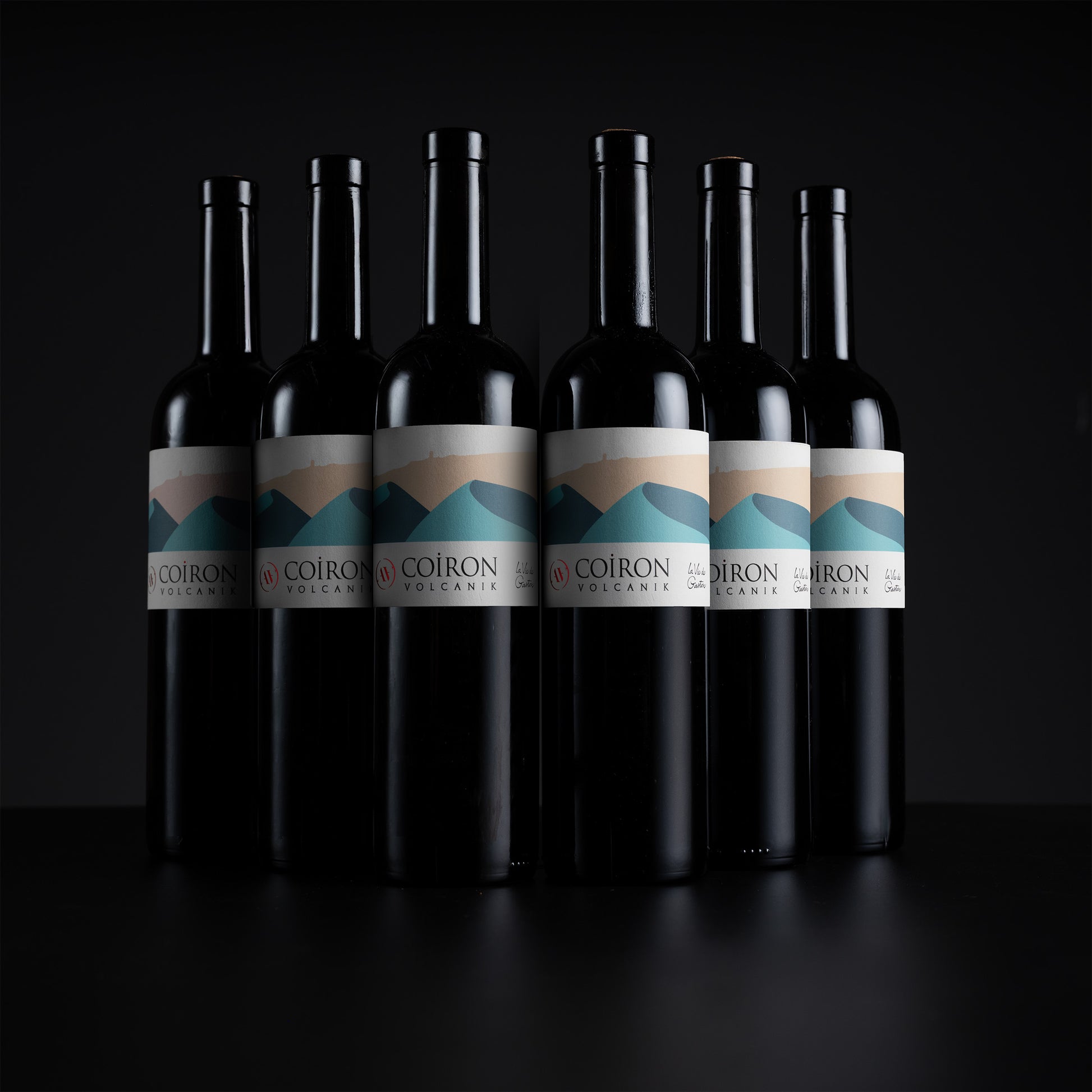 Six dark wine bottles with white labels featuring a blue and beige mountain graphic, arranged in a row against a black background. The label text reads 'COIRON OREGON'.