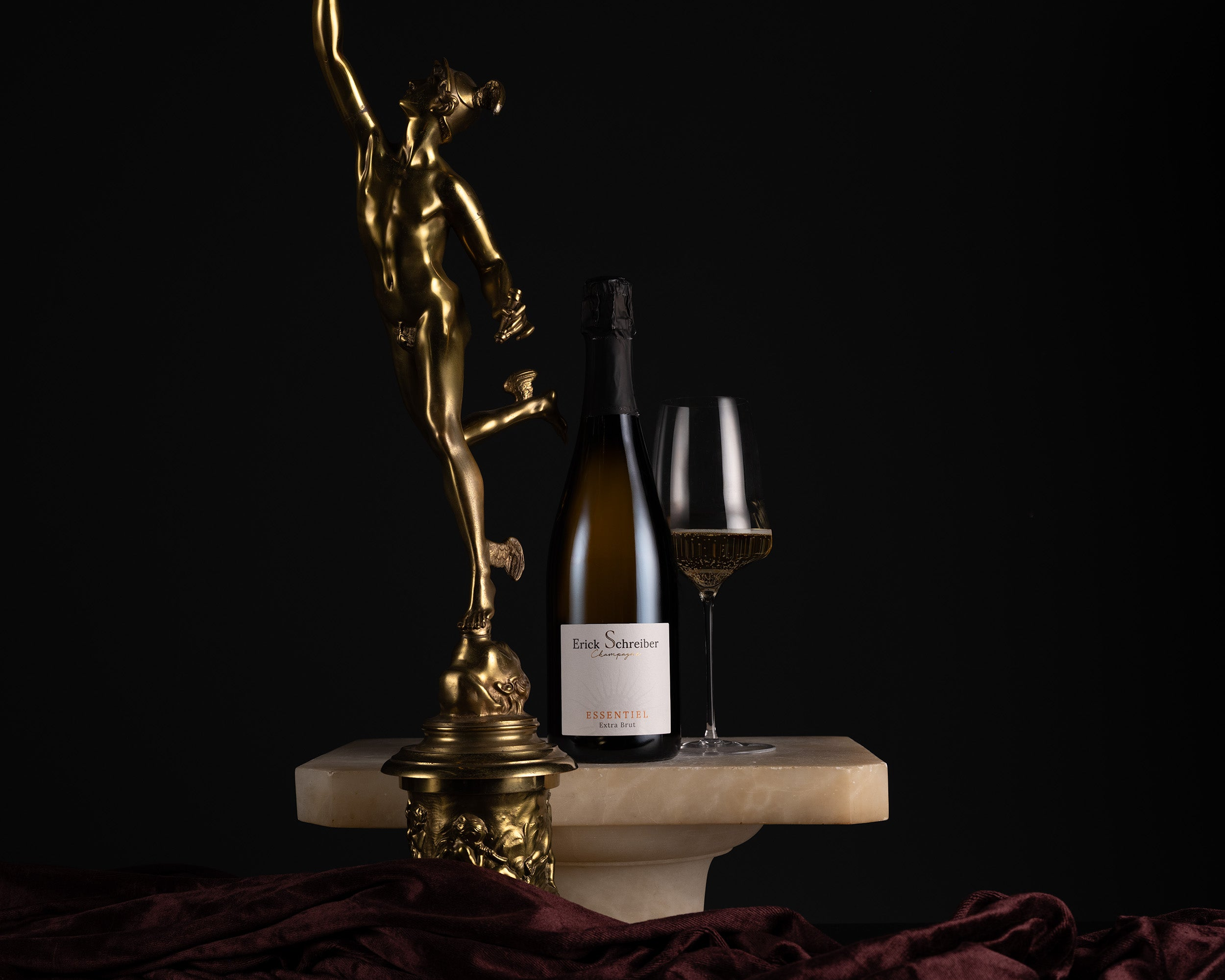 A bottle of Enoch Sutherland sparkling wine and a filled wine glass are styled on a white marble pedestal next to a gold statue against a black background.