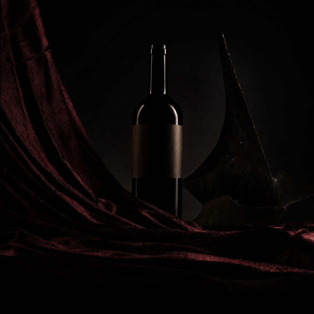 A dark wine bottle with a blank label stands centered against a black background, draped with deep burgundy velvet fabric and a dark sculptural object at its side.