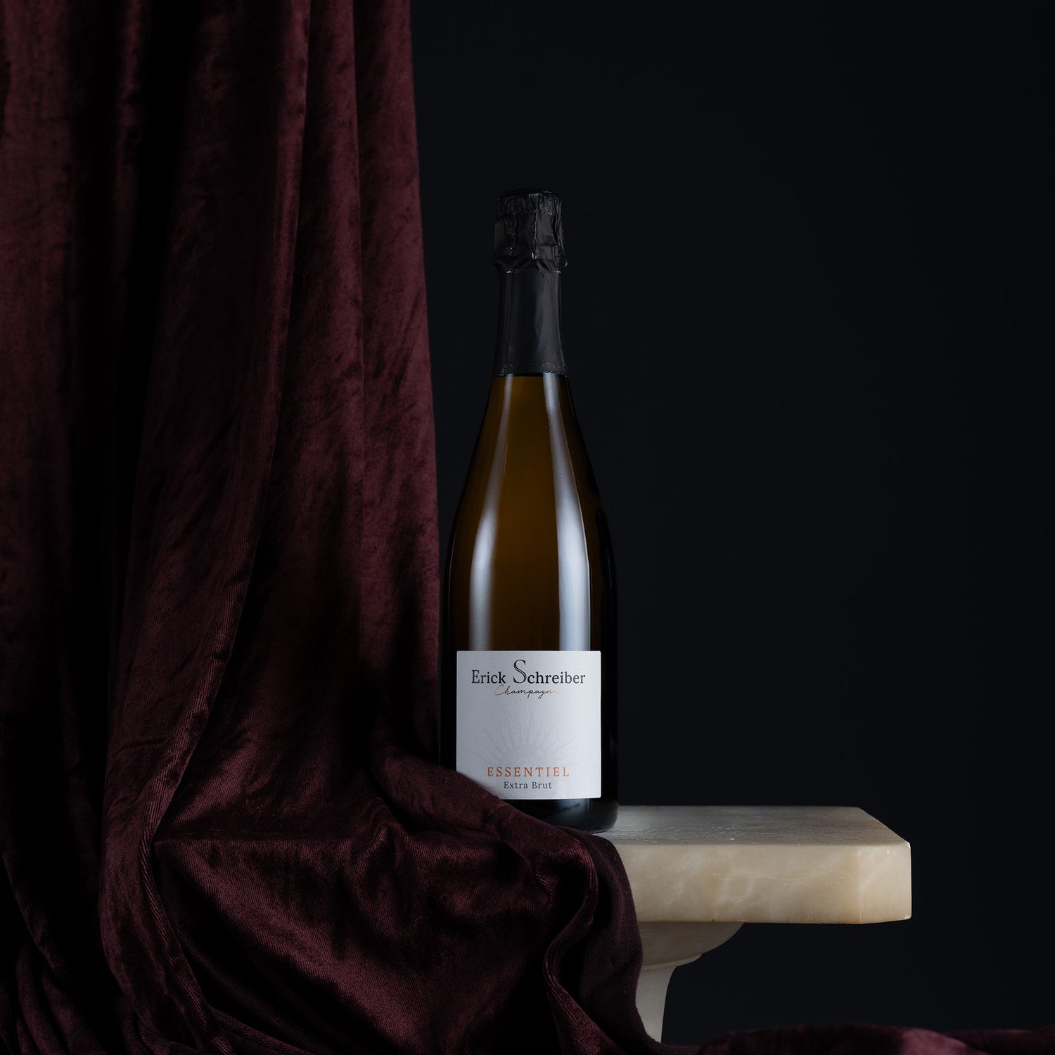 A bottle of Erich Schreiner Lavender & Rose sparkling wine with a white label, styled on a marble ledge next to a dark burgundy velvet drape against a black background.