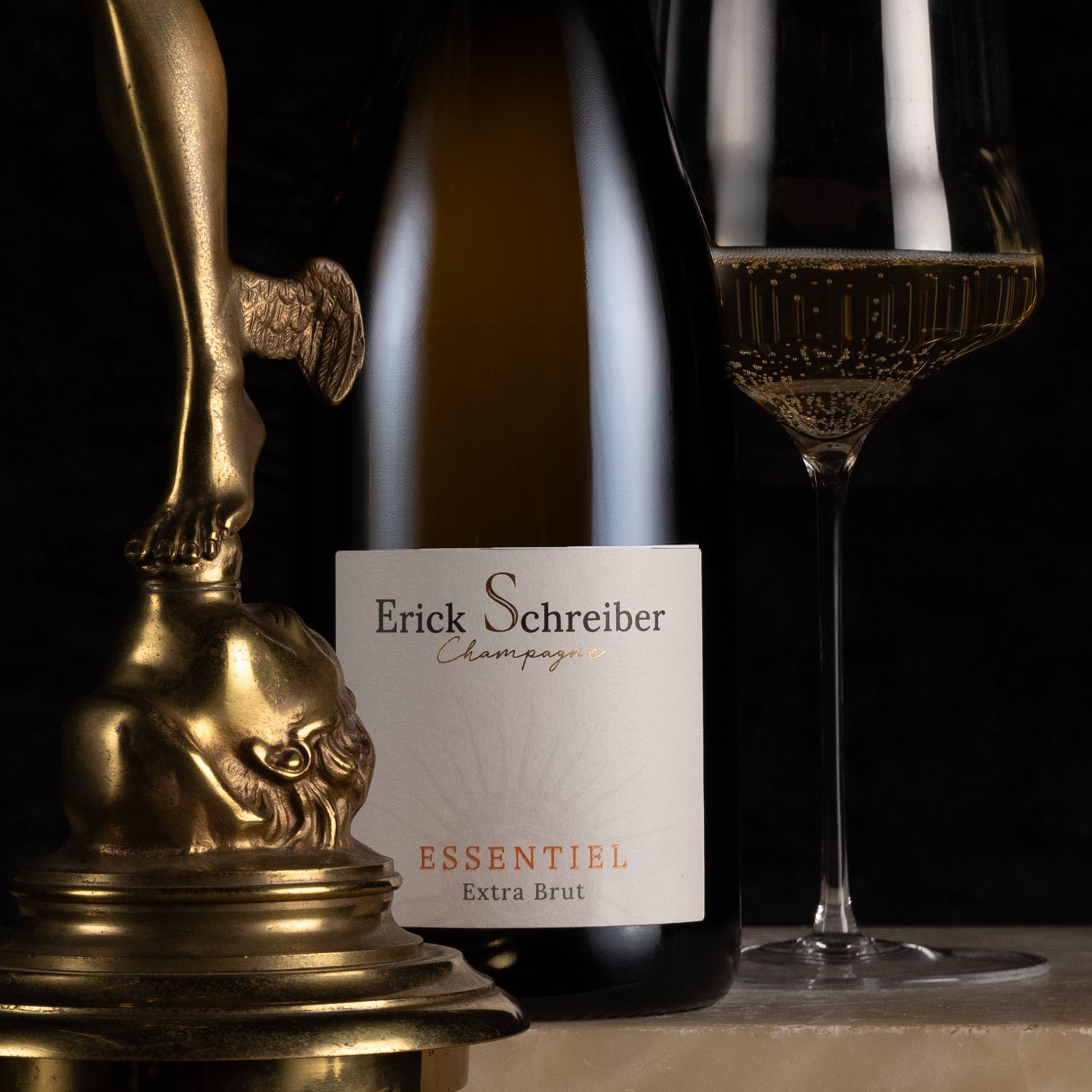 A bottle of Erick Schreiber Essentiel Extra Brut champagne stands next to a filled wine glass and a decorative gold sculpture against a dark background.