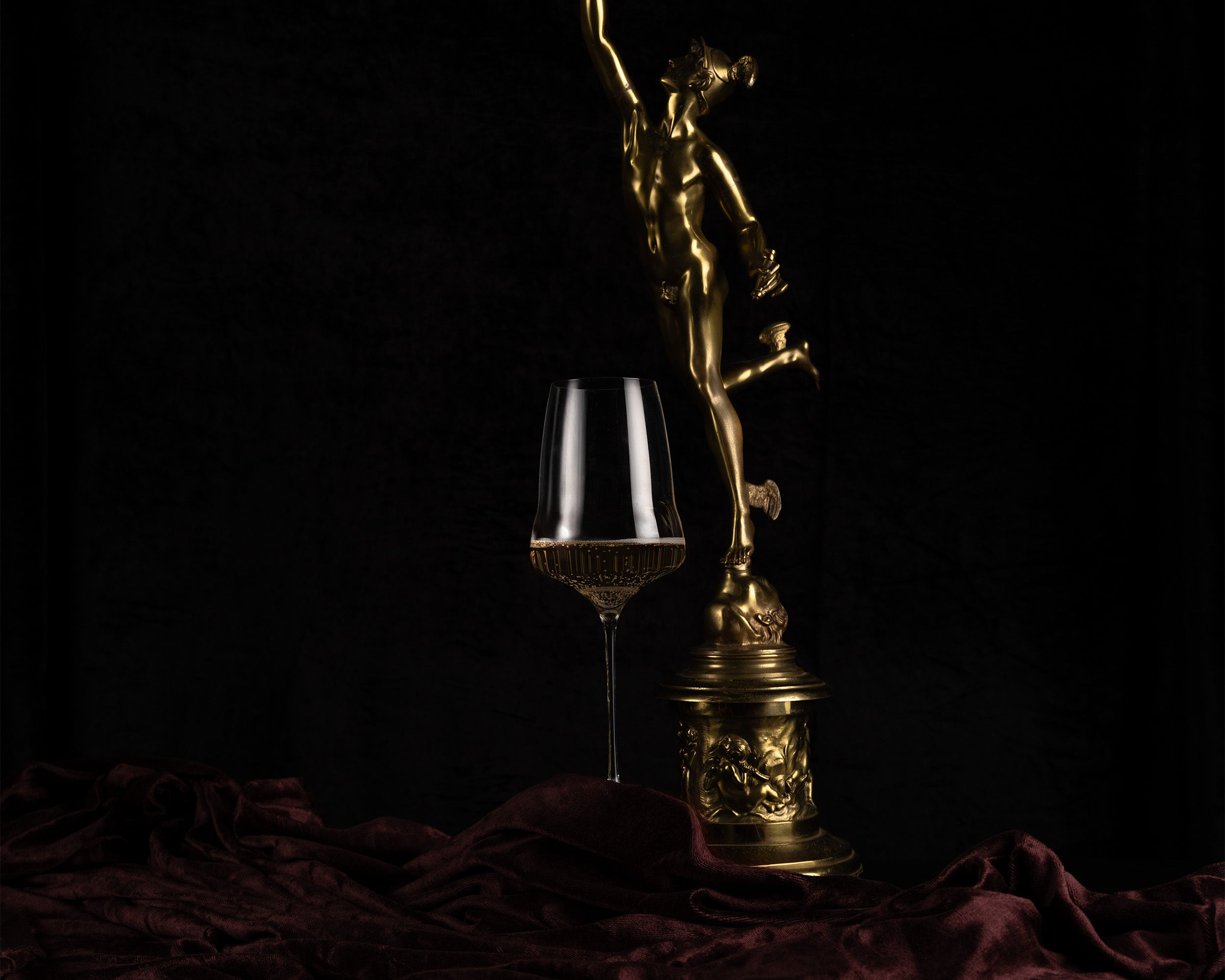 A gold-toned decorative figurine on an ornate pedestal stands beside a clear wine glass partially filled with red wine, set against a black background with draped burgundy fabric at the base.