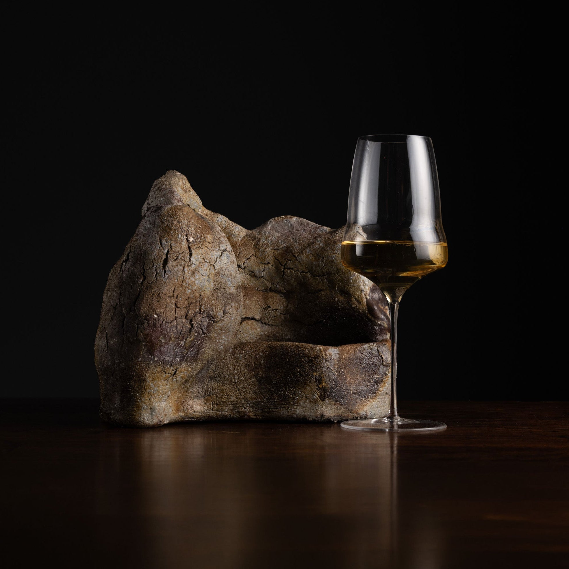 A clear wine glass partially filled with white wine sits on a dark wooden table beside a large, rough, stone-like decorative object against a black background.