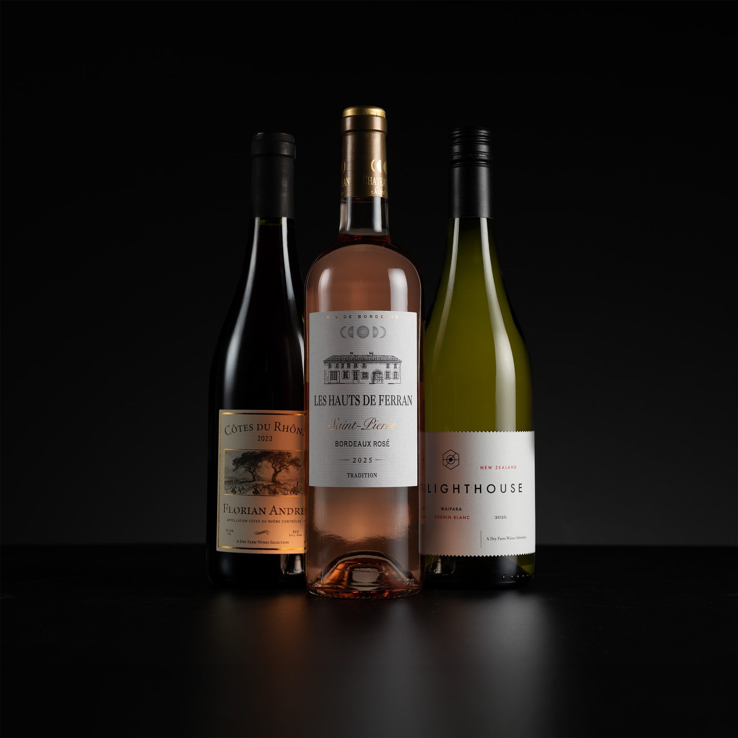 Three wine bottles arranged together on a black surface against a black background: a dark red bottle on the left, a rosé wine bottle in the center, and a green wine bottle on the right.