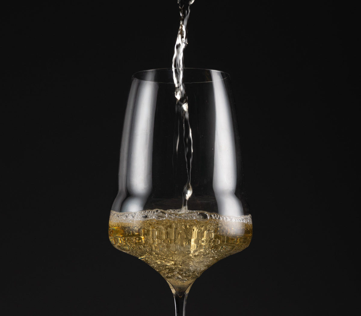 A clear stemmed wine glass with a wide bowl, shown on a black background with a stream of white wine being poured into it.