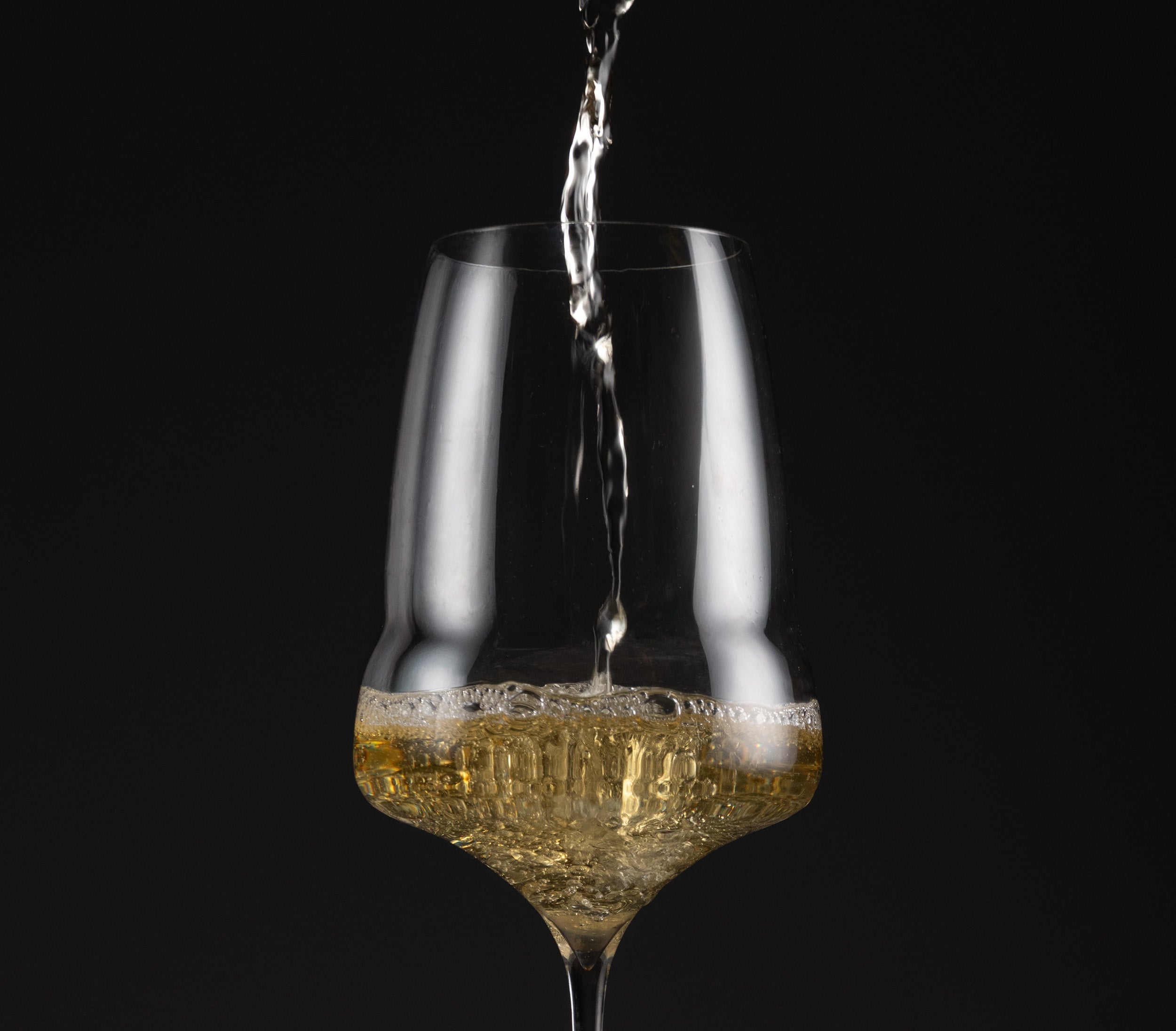A clear stemmed wine glass with a wide bowl, shown on a black background with a stream of white wine being poured into it.