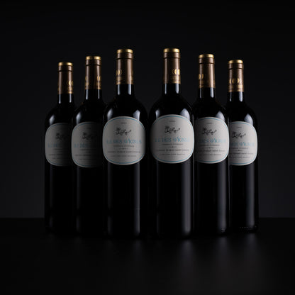 Six dark wine bottles with white labels and gold foil caps, arranged in a row against a black background. The center bottle is in sharp focus, with the others softly blurred in the background.