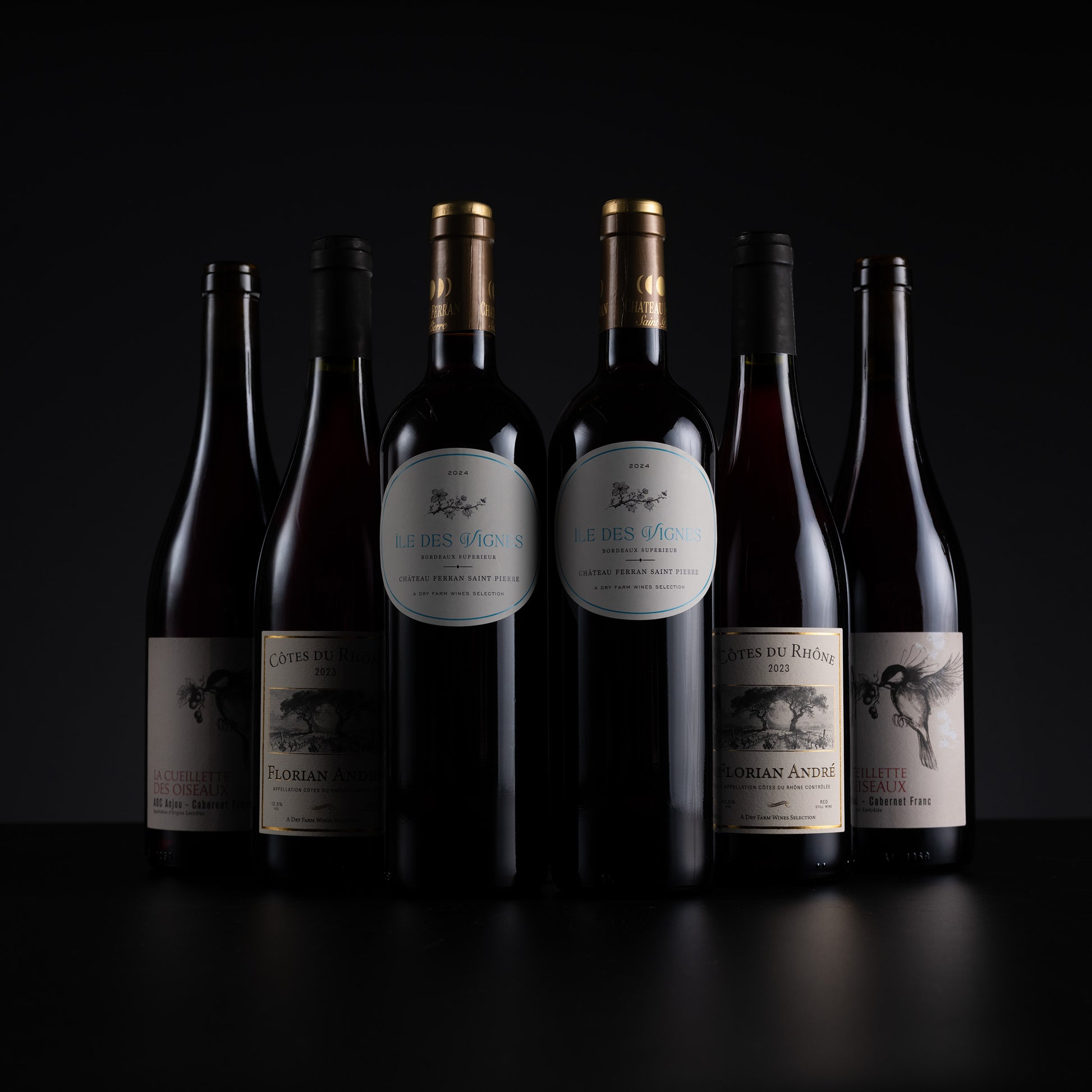 Six dark glass wine bottles with cream and gold labels arranged in a row against a black background. The center bottles have prominent white oval labels with blue text, while the side bottles have darker, less legible labels.