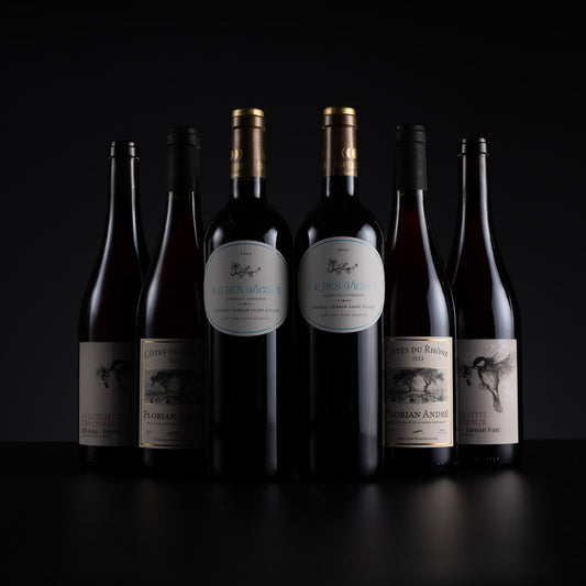 Six dark glass wine bottles with cream and gold labels arranged in a row against a black background. The center bottles have prominent white oval labels with blue text, while the side bottles have darker, less legible labels.