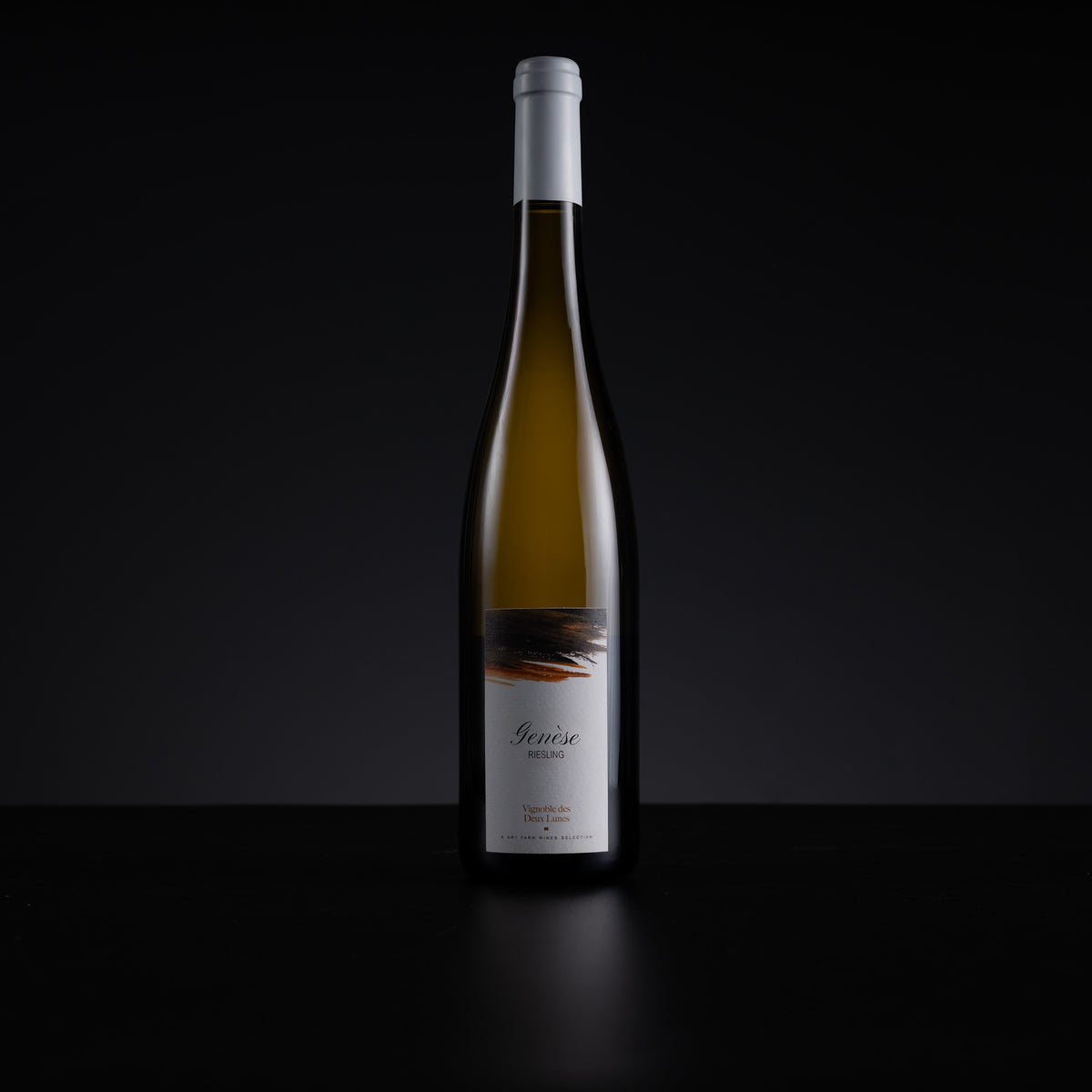 A single tall wine bottle with a white capsule and white label stands centered against a dark black background on a black surface. The bottle contains a pale golden liquid, and the label has a small abstract dark graphic near the top.