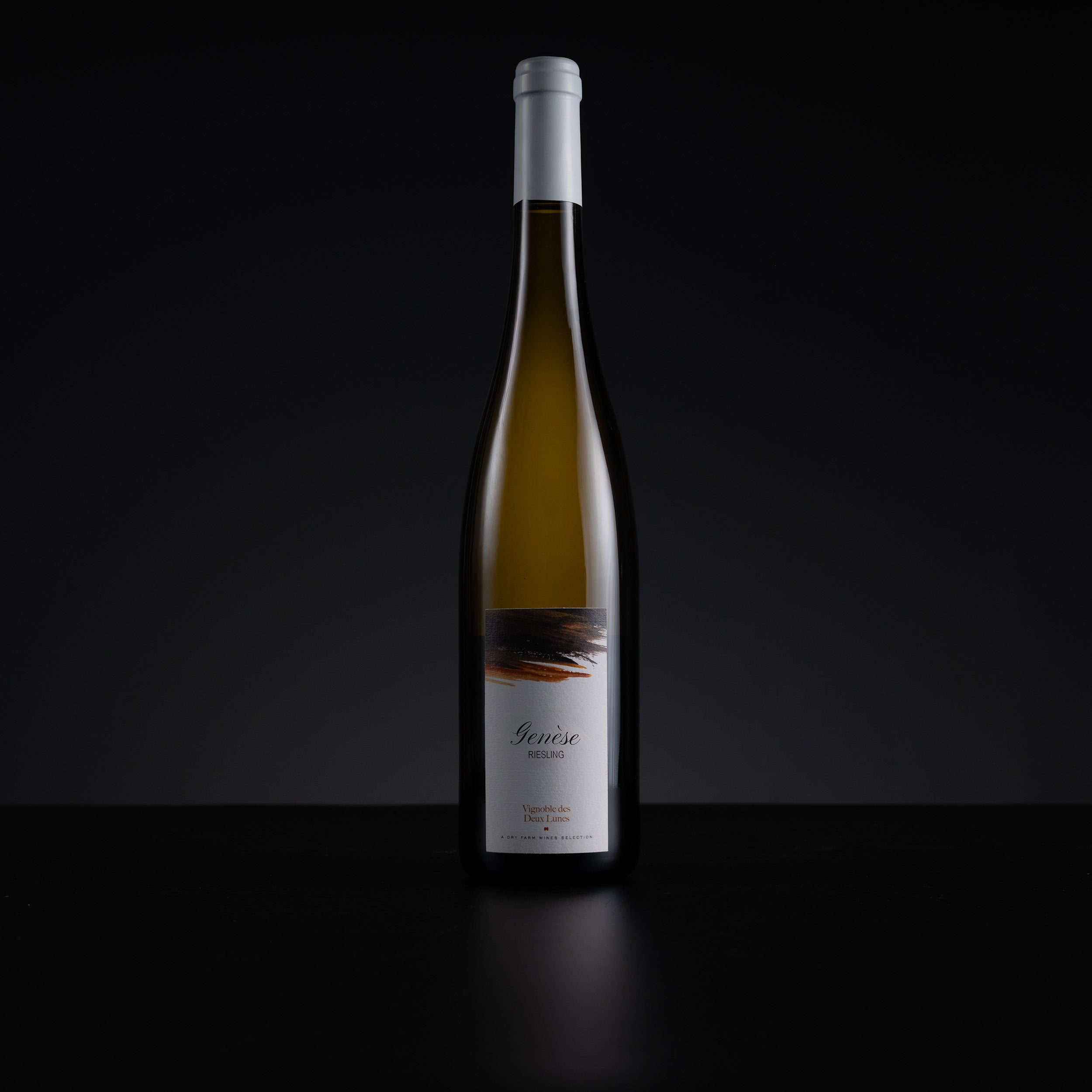 A single tall wine bottle with a white capsule and white label stands centered against a dark black background on a black surface. The bottle contains a pale golden liquid, and the label has a small abstract dark graphic near the top.