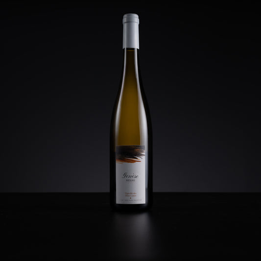 A single tall wine bottle with a white capsule and white label stands centered against a dark black background on a black surface. The bottle contains a pale golden liquid, and the label has a small abstract dark graphic near the top.