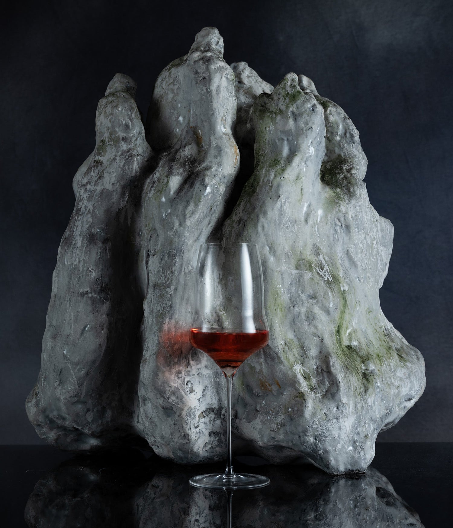 A clear wine glass with a small amount of red liquid stands in front of a large, rough, sculptural gray stone or rock formation on a dark reflective surface against a dark background.