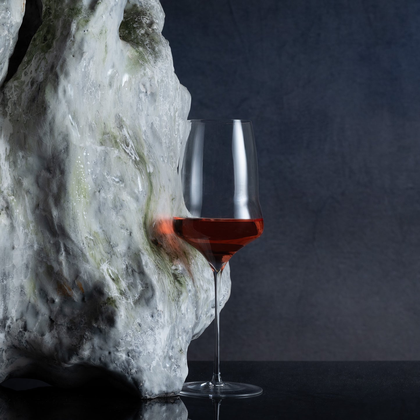 A clear wine glass with a wide bowl, partially filled with red wine, standing beside a large textured white stone on a dark reflective surface against a dark gray background.