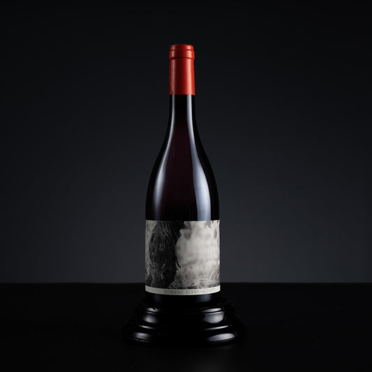 A dark glass wine bottle with a red foil capsule and a black-and-white label, displayed upright on a black pedestal against a dark gradient background.