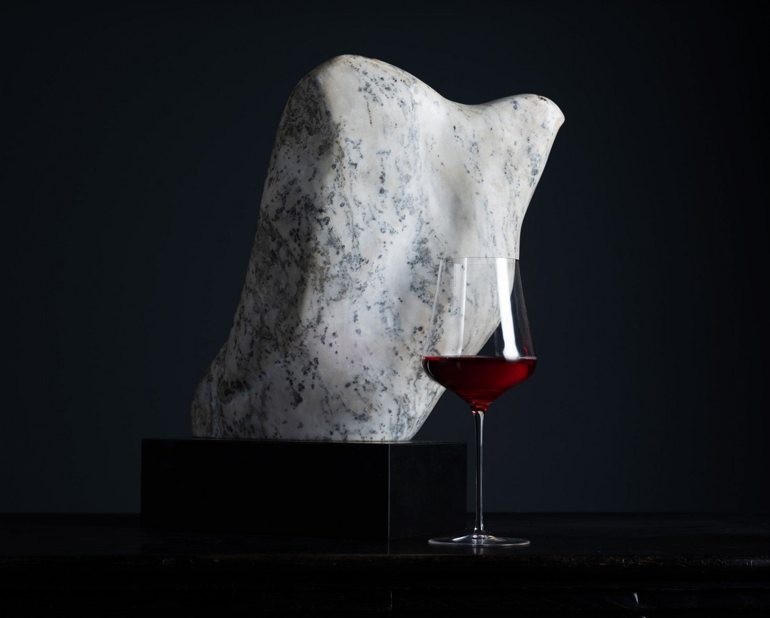 A sculptural white stone or marble object with dark speckling stands on a black pedestal, with a stemmed wine glass holding red wine placed in front, against a very dark studio background.