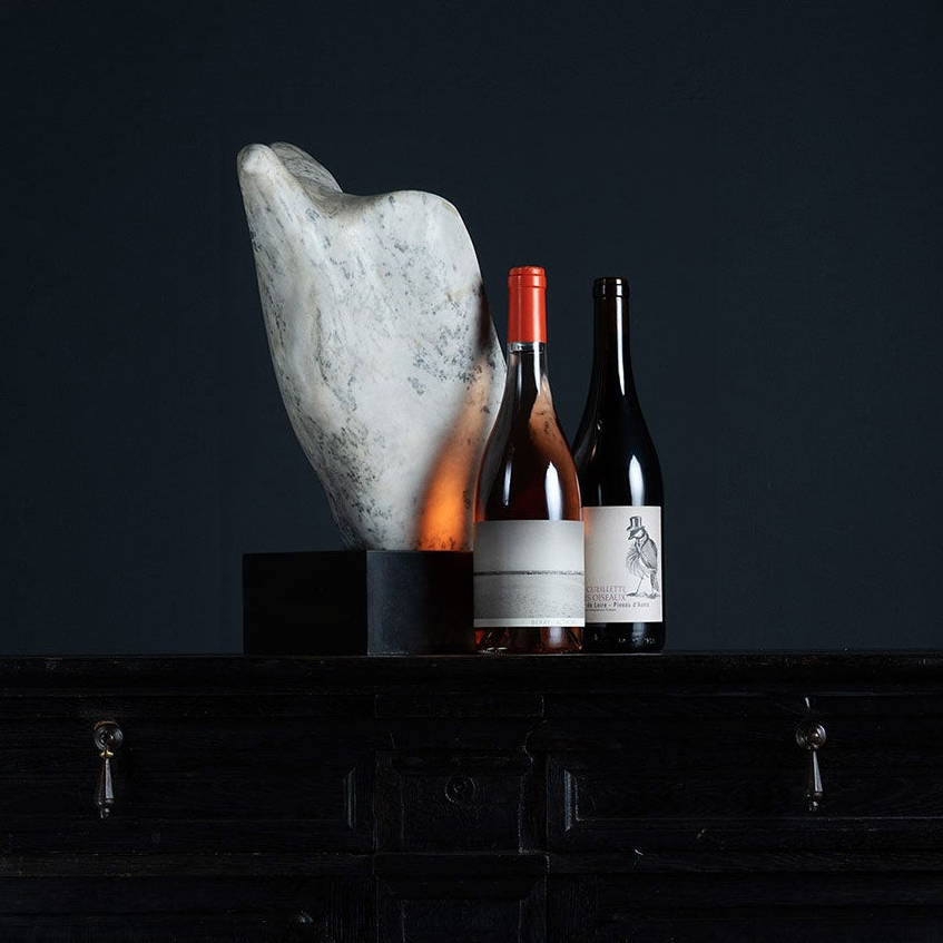 Two wine bottles with white labels and red/orange caps stand on a black wooden cabinet against a dark navy background, next to a large white stone sculpture on a black base.
