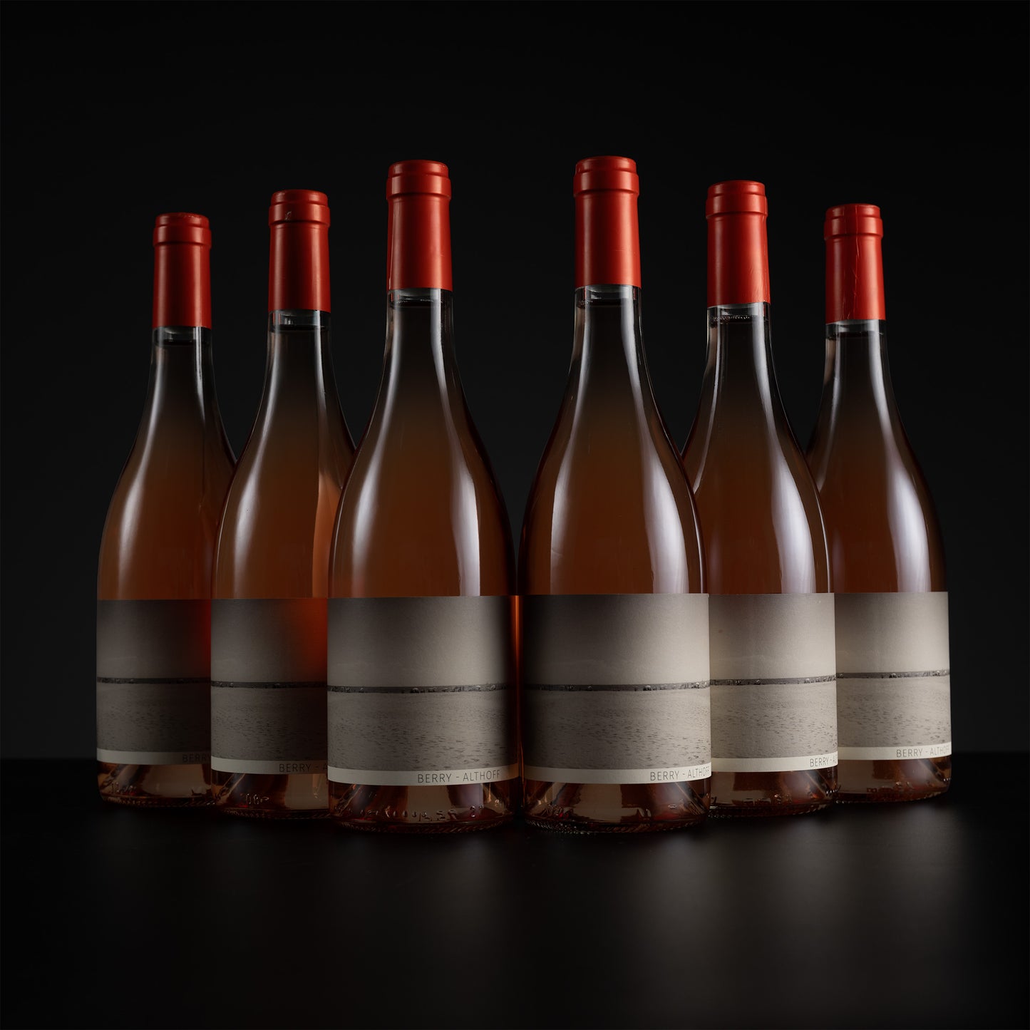 Six wine bottles with red foil caps and blank gray labels, arranged in a row on a dark reflective surface against a black background.