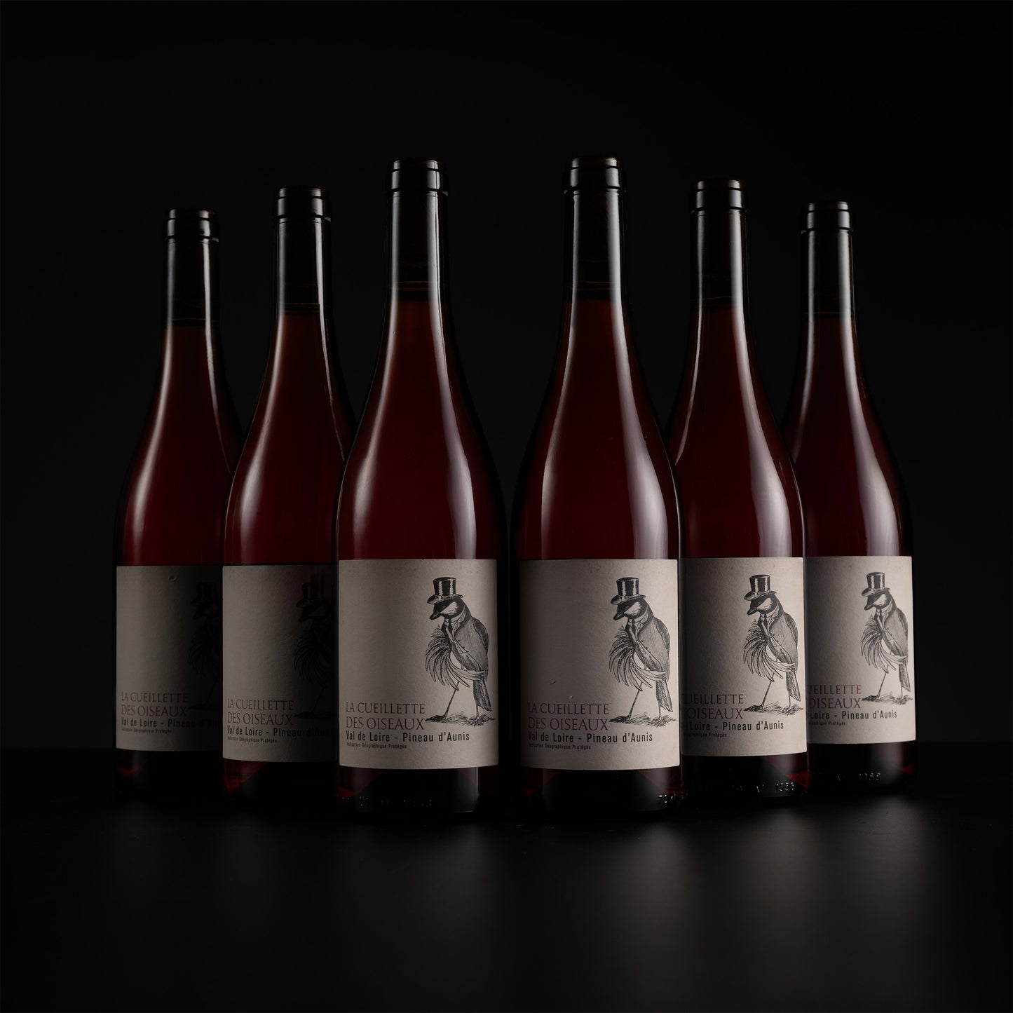 Six dark glass wine bottles with cream labels and black capsules, arranged in a row against a black background. The central bottle is in sharp focus, with the others softly blurred behind it.