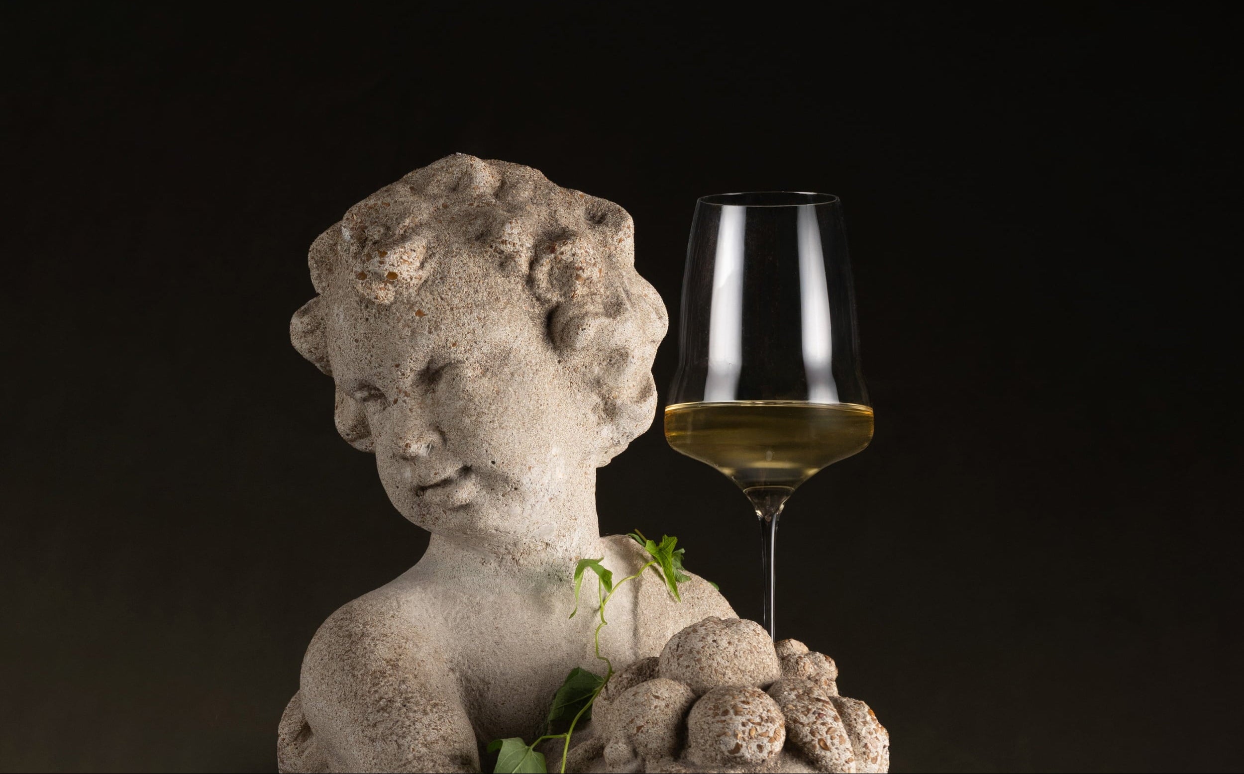 A clear stemmed wine glass filled with white wine stands beside a small stone cherub statue holding grapes, set against a dark black background.