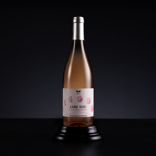 A bottle of Rosé wine displayed on a black stand against a dark studio background. The bottle contains a pale pinkish-rose liquid and features a white label with pink floral illustrations and the text 'LUNE ROSE' and 'CÔTE D'EAU DE PROVENCE'.