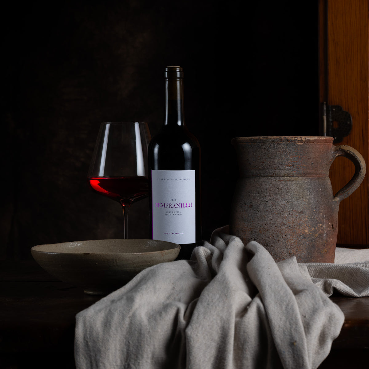A dark wine bottle with a white label reading 'VIGNETTE DI PIANELLO' stands beside a partially filled wine glass, a ceramic bowl, and a rustic ceramic pitcher on a draped cloth against a dark background.