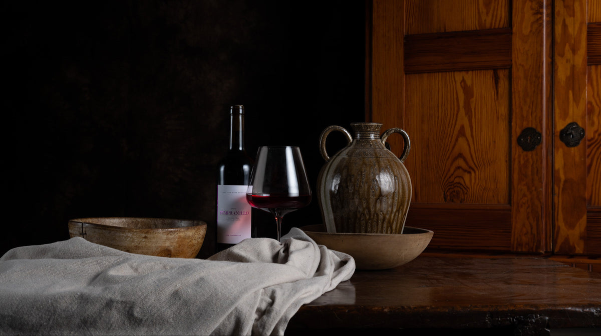 A rustic still life scene with a dark wine bottle, a filled wine glass, a ceramic jug, wooden bowls, and a draped cloth on a wooden table against a dark background and wood-paneled wall.
