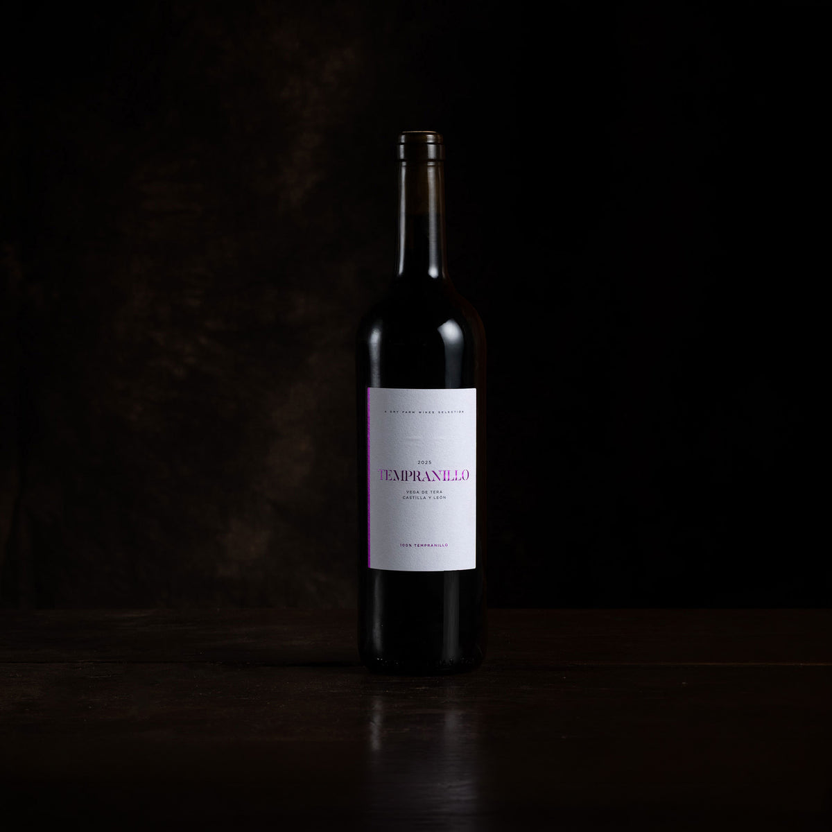 A single dark wine bottle with a white label featuring pink text, standing on a dark wooden surface against a black background.
