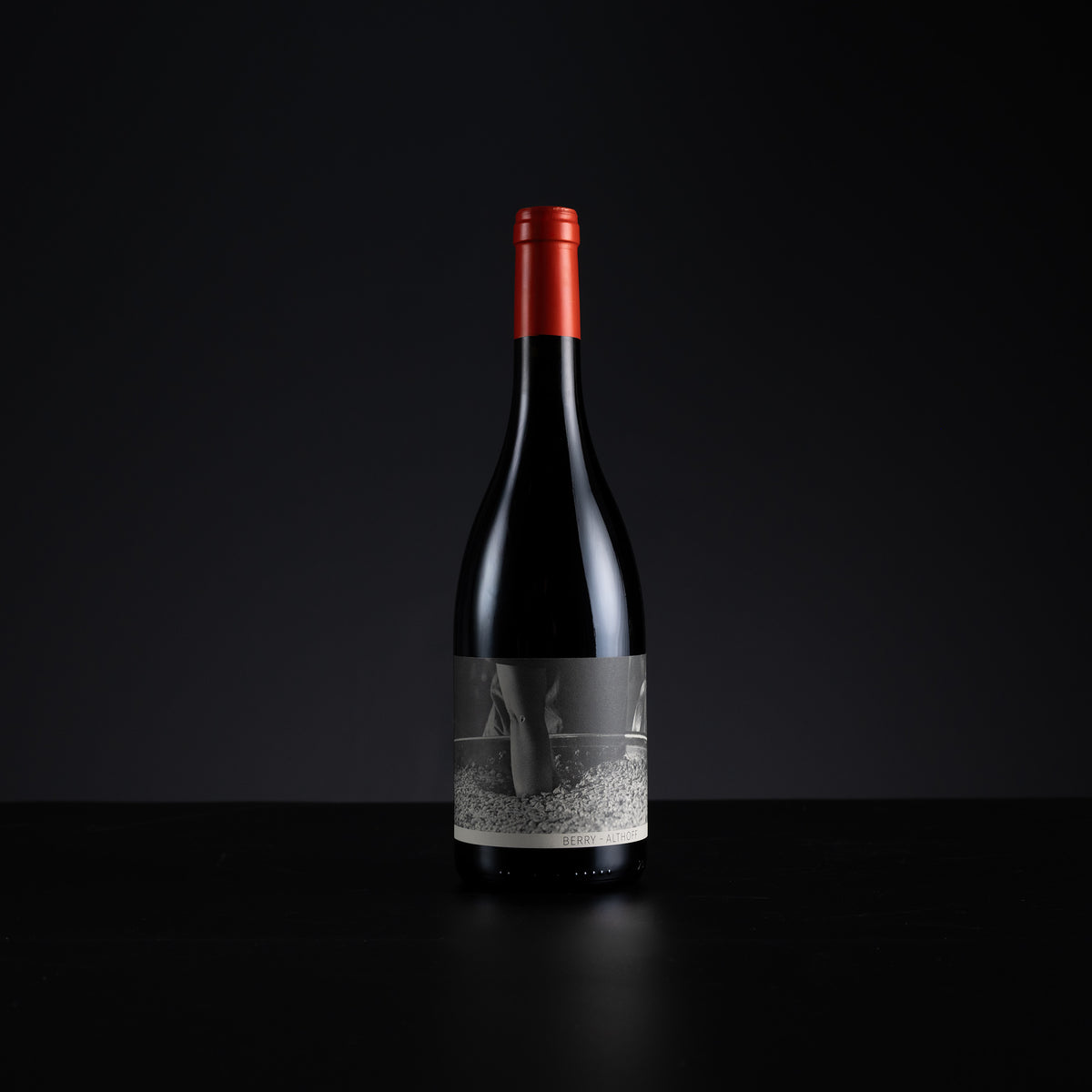 A single dark wine bottle with a red foil capsule and a grayscale label, standing centered on a black surface against a dark background.