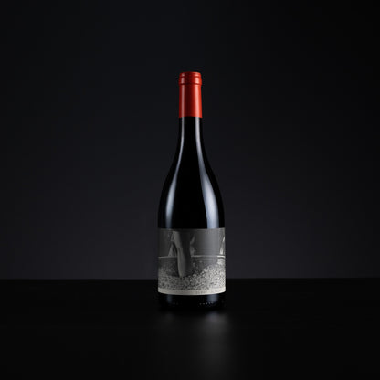 A single dark wine bottle with a red foil capsule and a grayscale label, standing centered on a black surface against a dark background.
