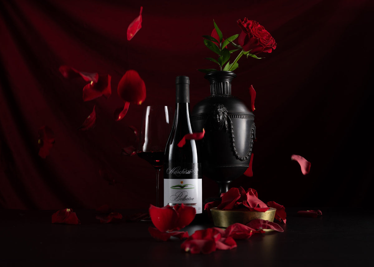 A bottle of red wine with a glass and a black vase holding red roses, styled with scattered red rose petals against a dark red draped background.