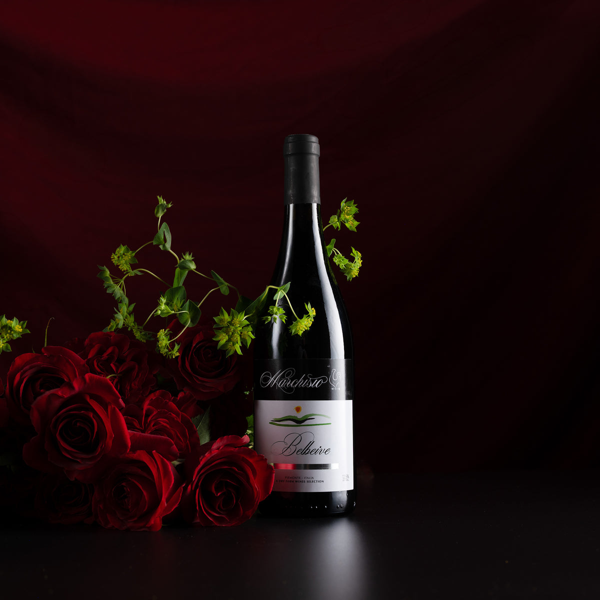 A dark wine bottle with a black cap and a white label reading 'Mondavi' and 'Believe', styled with red roses and green stems against a dark red draped background.