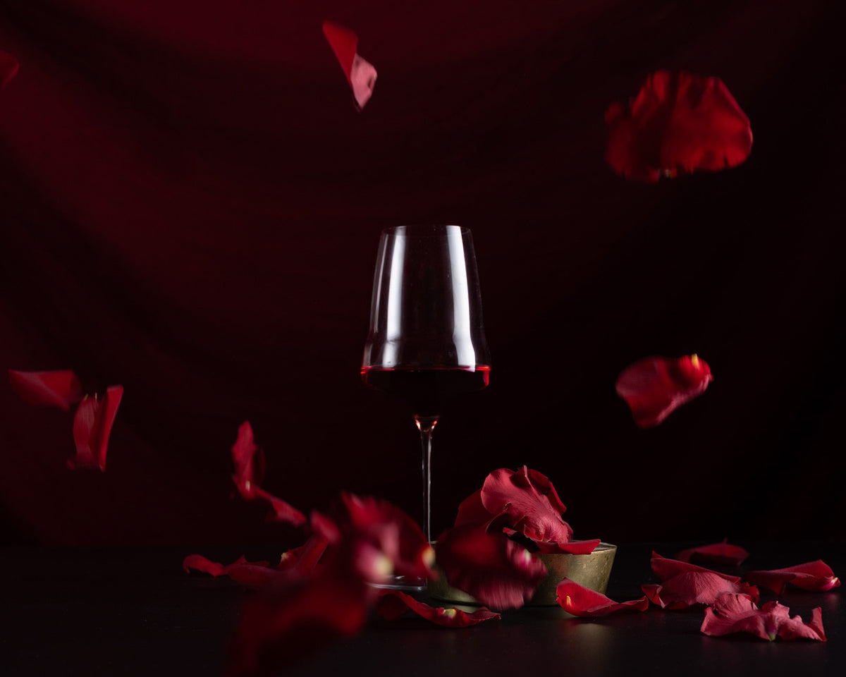 A single stemmed wine glass filled with red wine stands centered on a dark surface, surrounded by scattered and floating red rose petals against a deep red background.