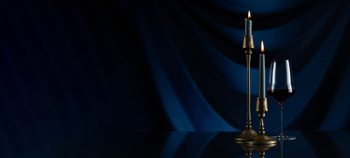 Two lit brass candlesticks and a wine glass with red wine on a reflective dark surface against a draped deep blue fabric backdrop.