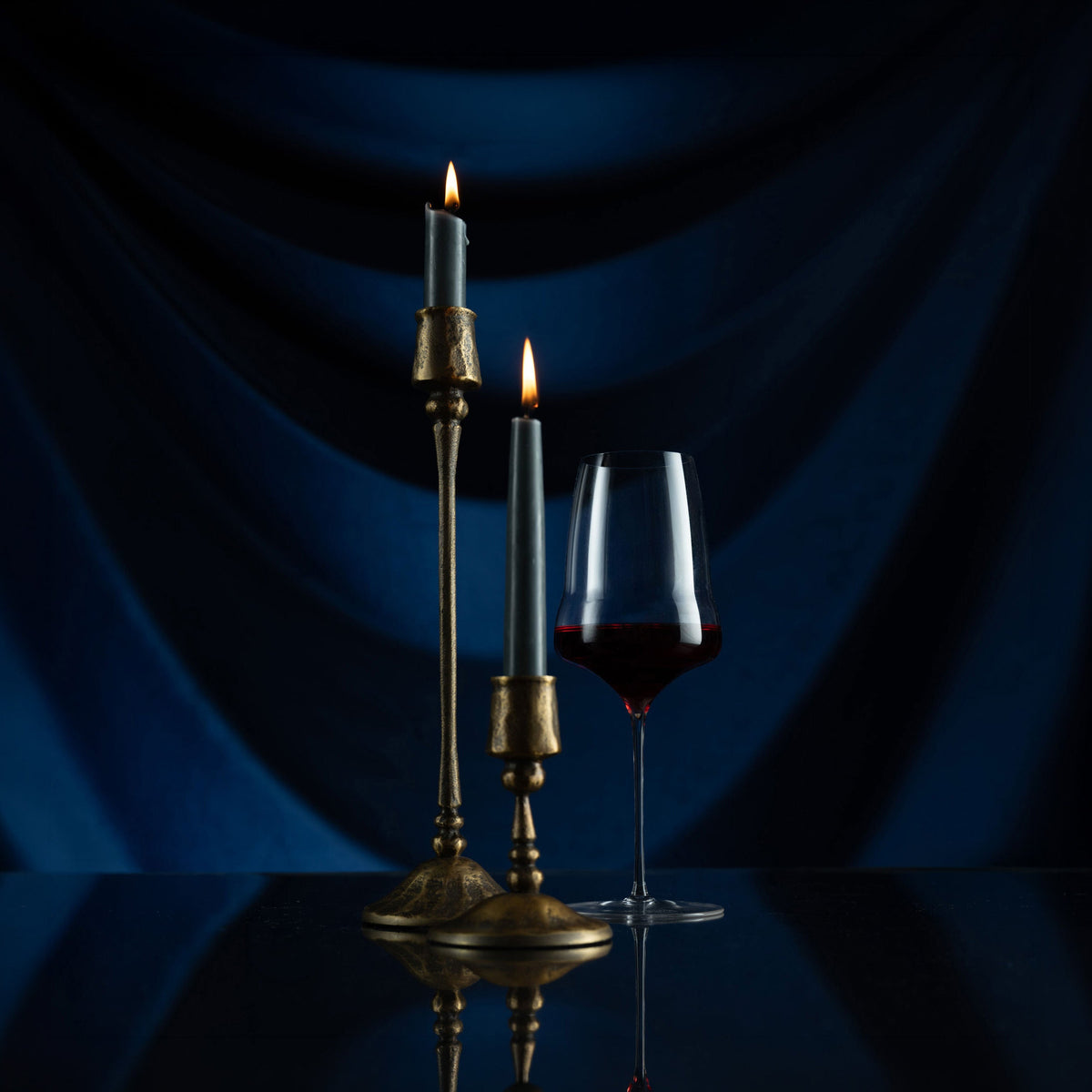 A dark moody still life with two lit taper candles in brass holders and a wine glass containing red wine, set against a draped deep blue fabric backdrop on a reflective black surface.