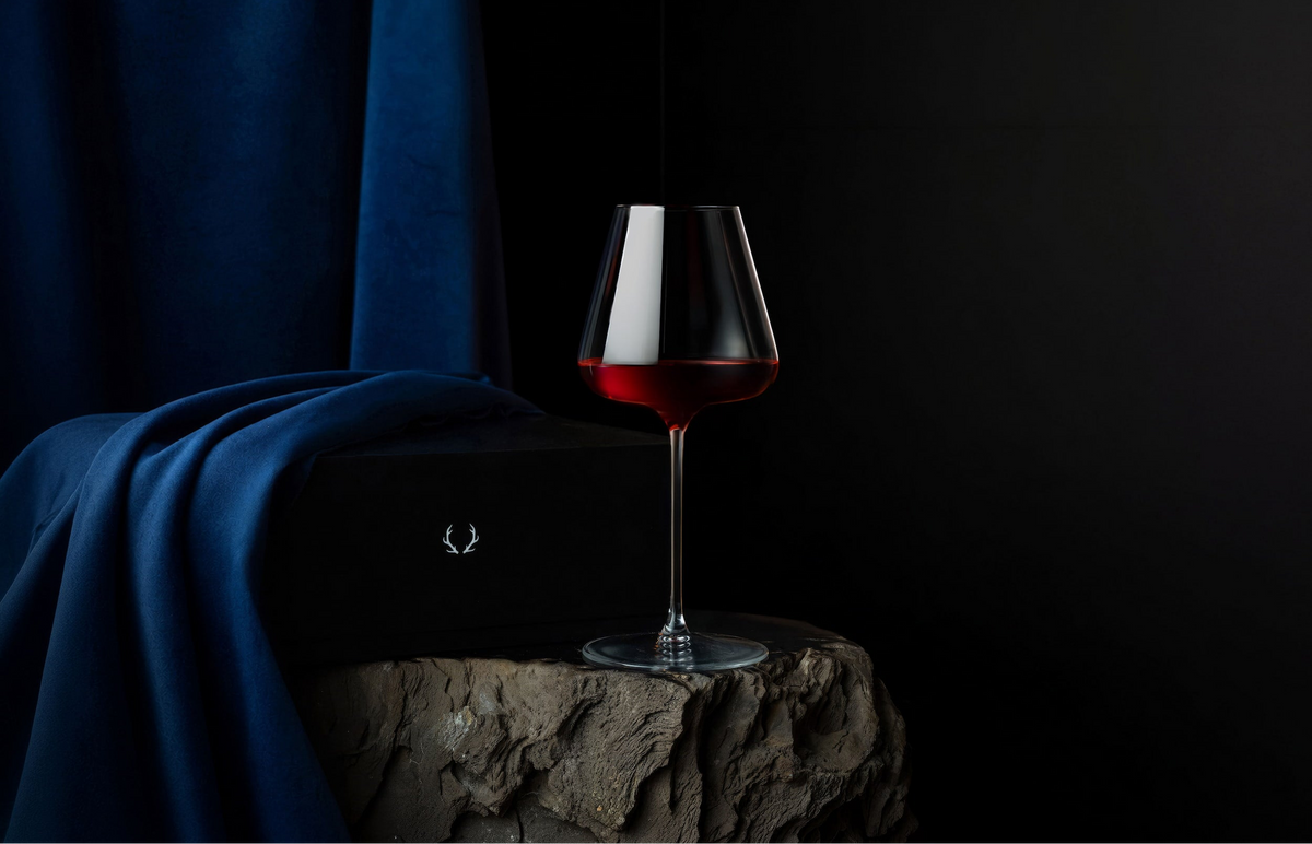 A clear wine glass with a wide bowl partially filled with red wine, set on a stone pedestal against a dark background with a draped blue fabric and a black box with a small silver laurel logo.