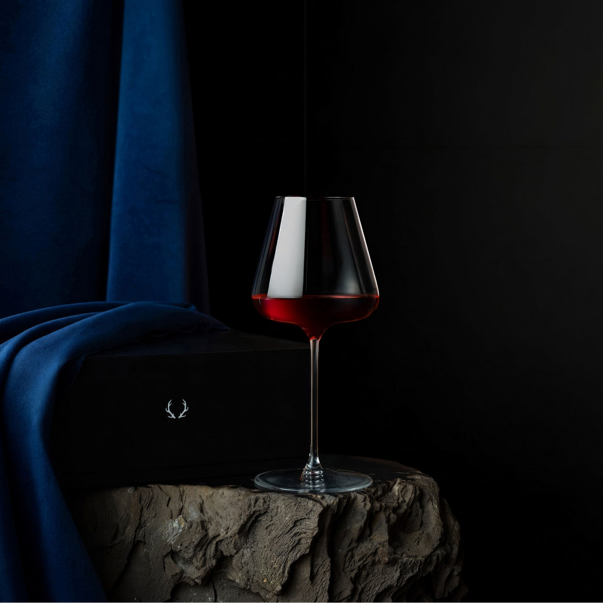 A stemmed wine glass partially filled with red wine, set on a rough stone pedestal against a dark background with a draped blue fabric and a black box featuring a small deer logo.