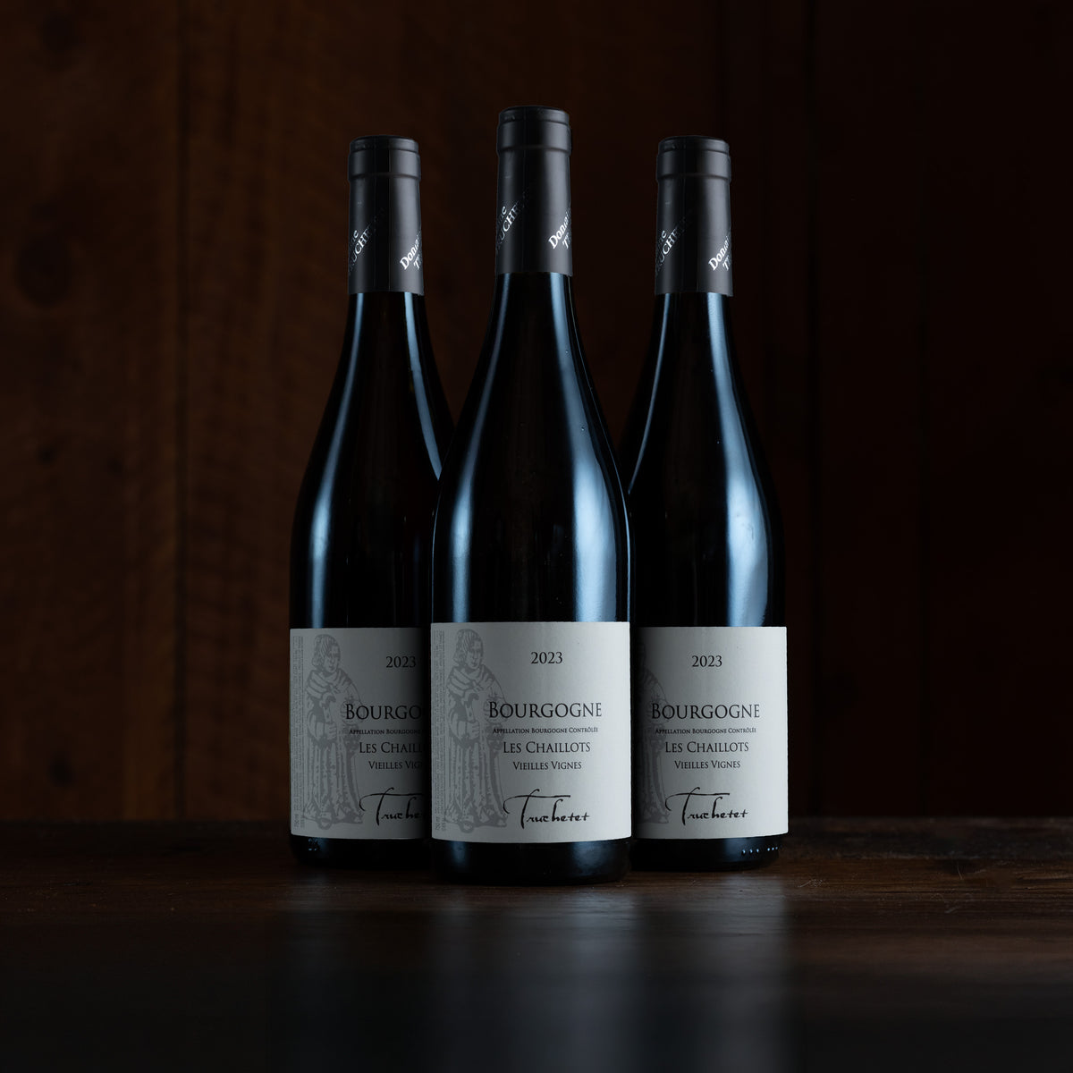 Three dark wine bottles with white labels and black capsules stand on a dark wooden surface against a dark wood background.