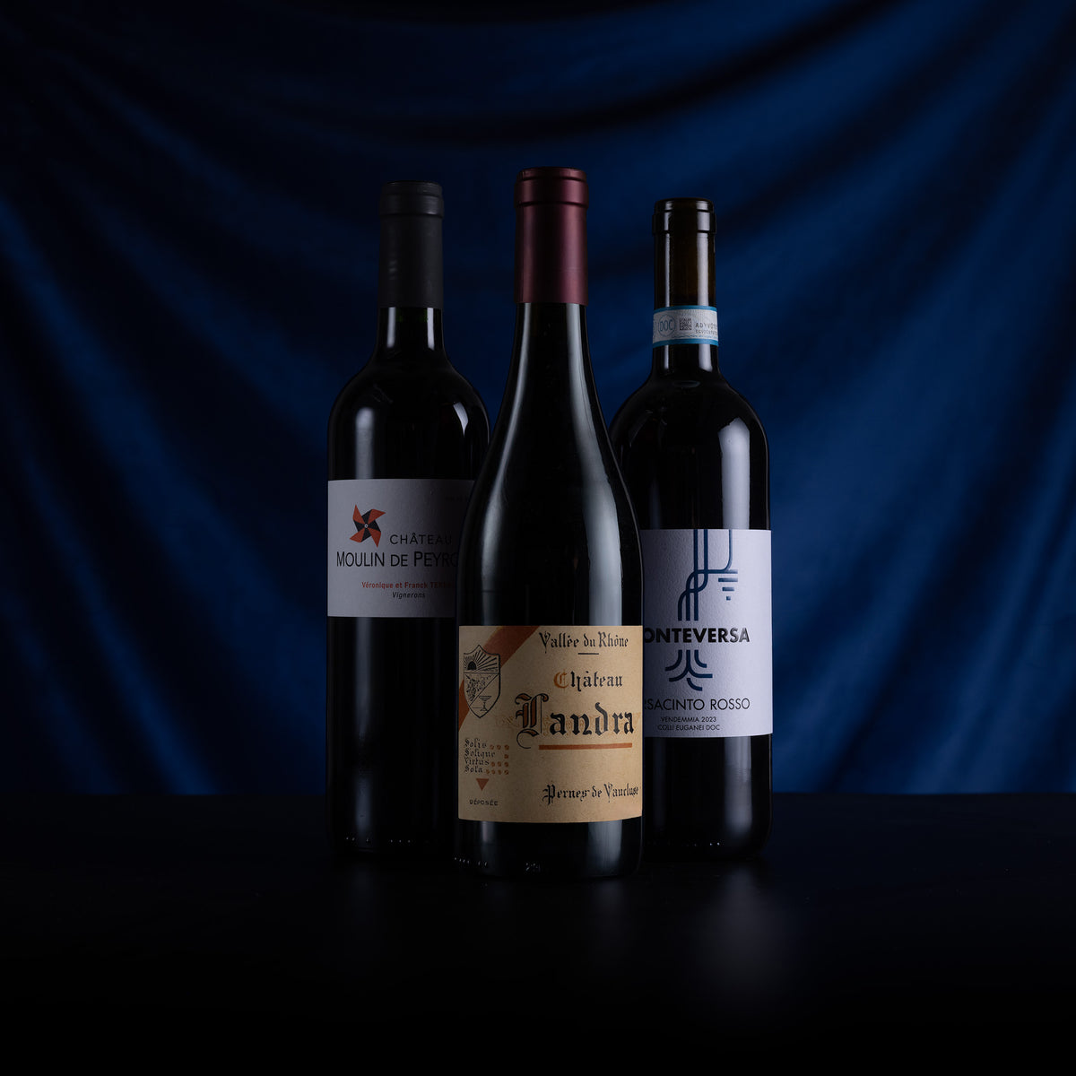 Three dark wine bottles arranged on a black surface against a draped dark blue background. The center bottle is upright and labeled 'Vall de Riu L'andra', flanked by two darker bottles with white and black labels.
