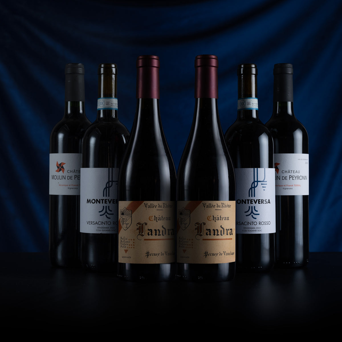 Six dark wine bottles from the Monteverga winery arranged in a row on a dark surface against a deep blue draped background. The bottles have black, burgundy, and dark red foil tops, and their white and beige labels display different wine varietals including Chardonnay, Sauvignon, and Merlot.