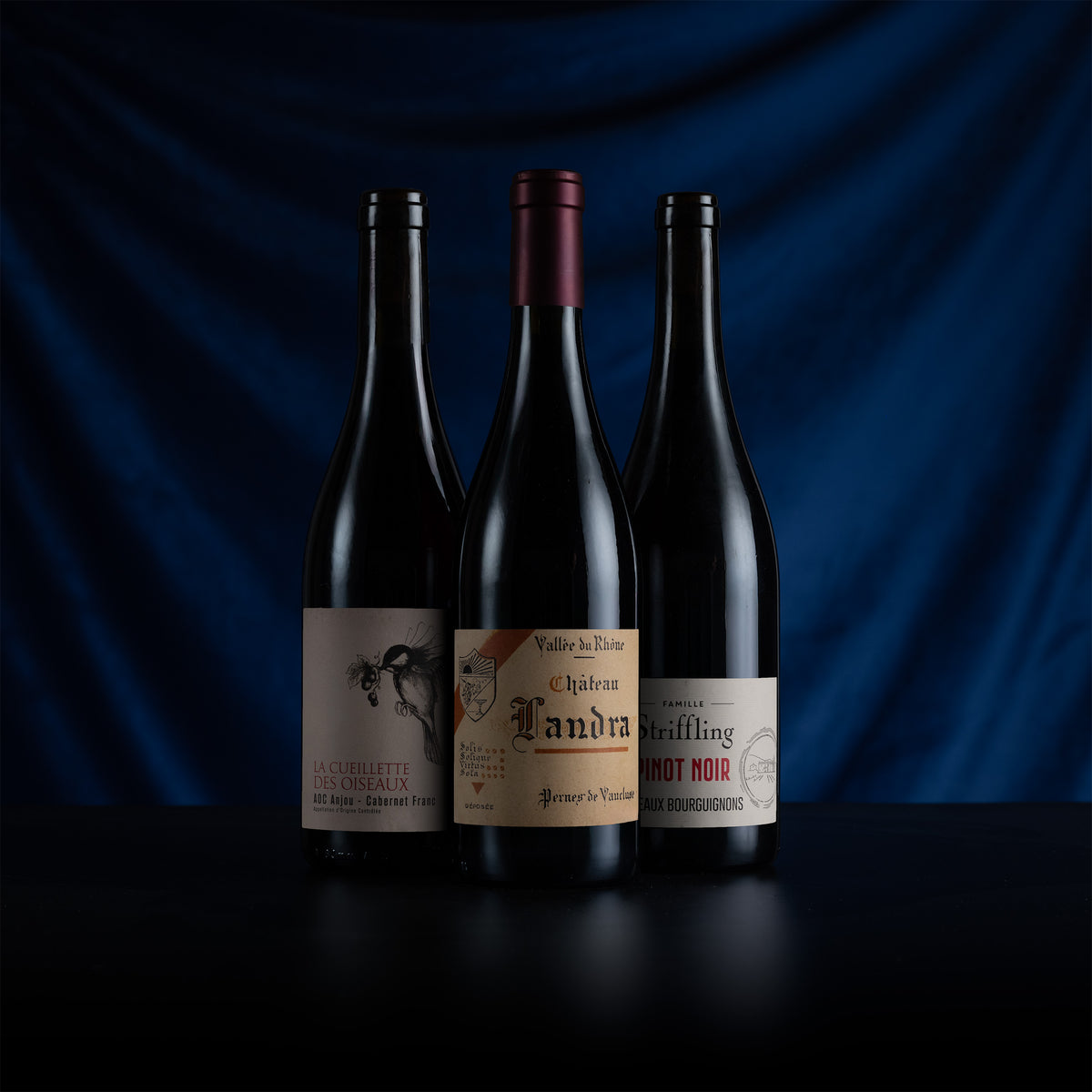 Three dark glass wine bottles standing upright on a black surface against a draped dark blue fabric background. The center bottle has a burgundy capsule and a beige label reading 'Château Landra'; the left bottle has a beige label reading 'La Châbleau des Vignerons'; the right bottle has a beige label reading 'Griffing'.