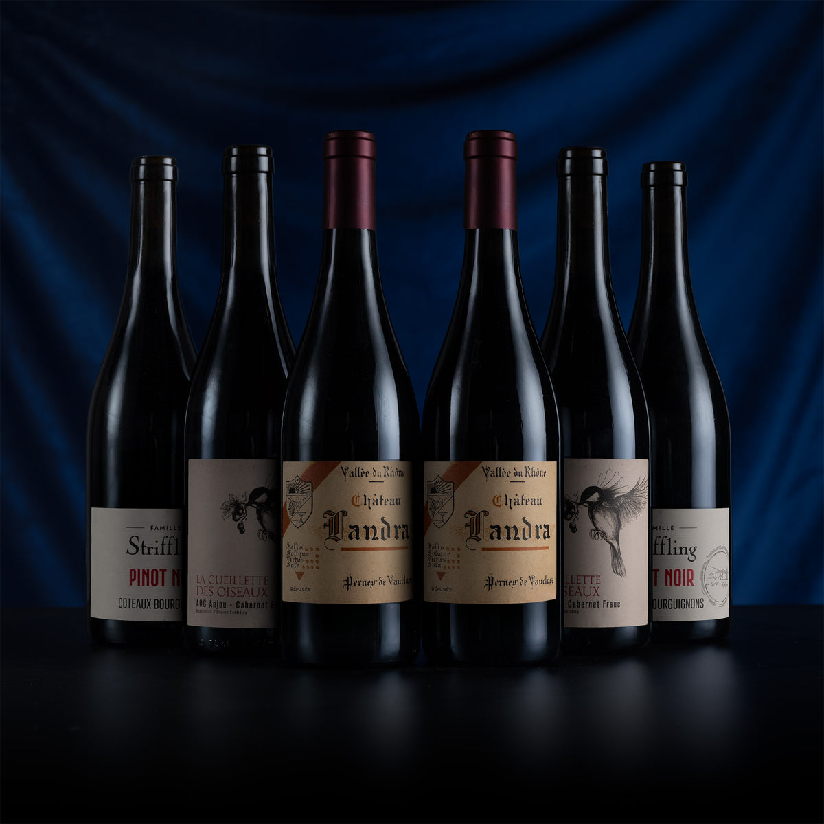 Six dark wine bottles arranged in a row on a black surface against a deep blue draped background. The center bottles show cream labels with black gothic text and illustrations, including Valdaine, Châbleau, and Andra.