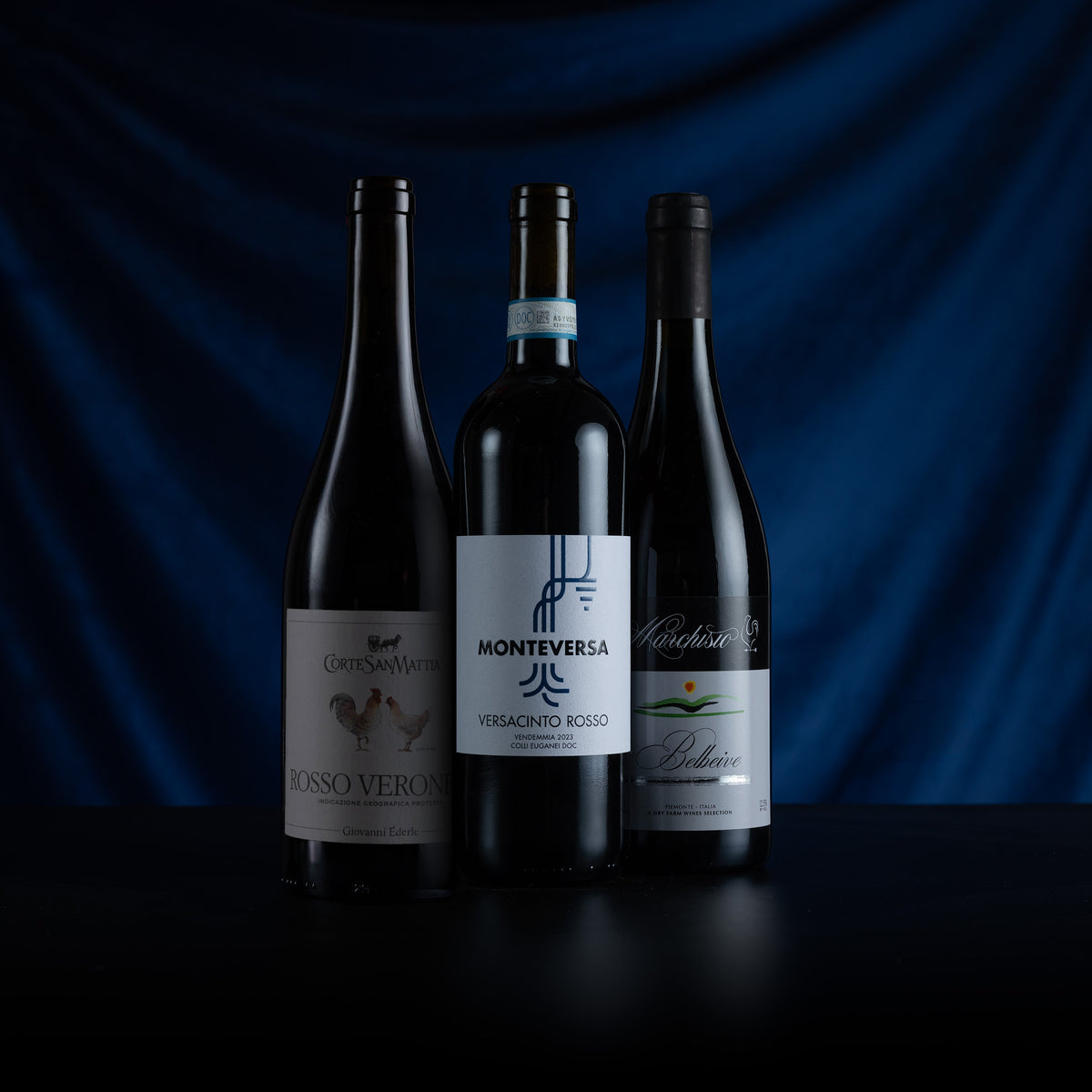 Three dark glass wine bottles standing on a black surface against a draped dark blue background. The center bottle has a white label reading Monteversa Veracino Rosso, flanked by two other dark bottles with cream and green labels.