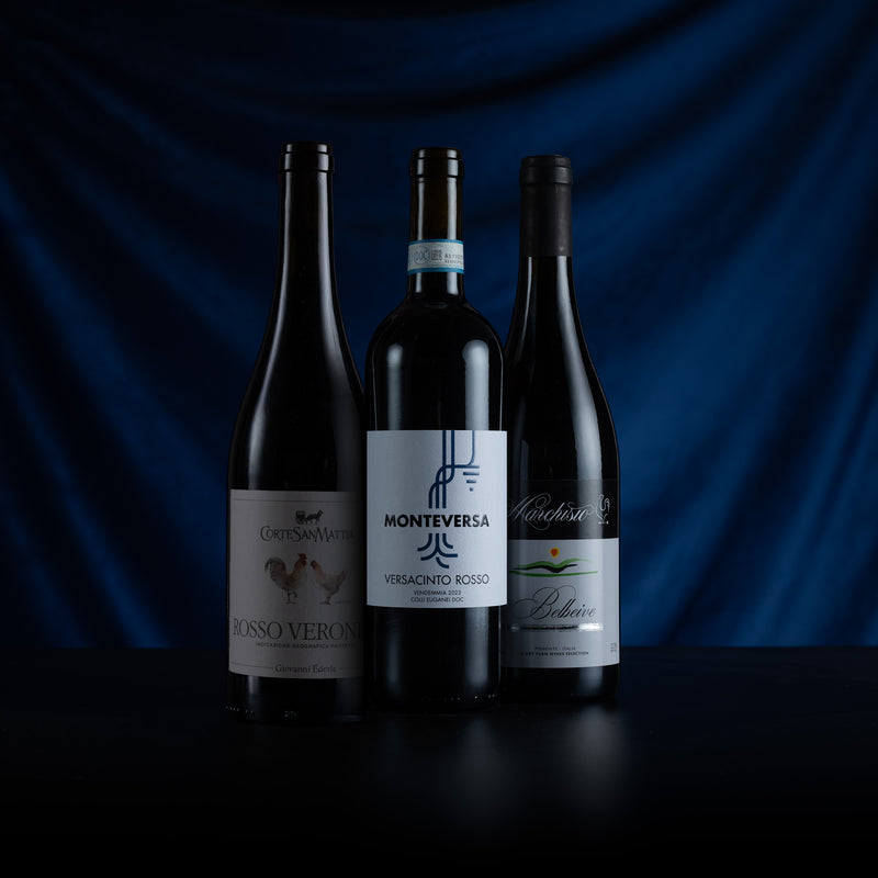 Three dark glass wine bottles standing on a black surface against a draped dark blue background. The center bottle has a white label reading Monteversa Veracino Rosso, flanked by two other dark bottles with cream and green labels.