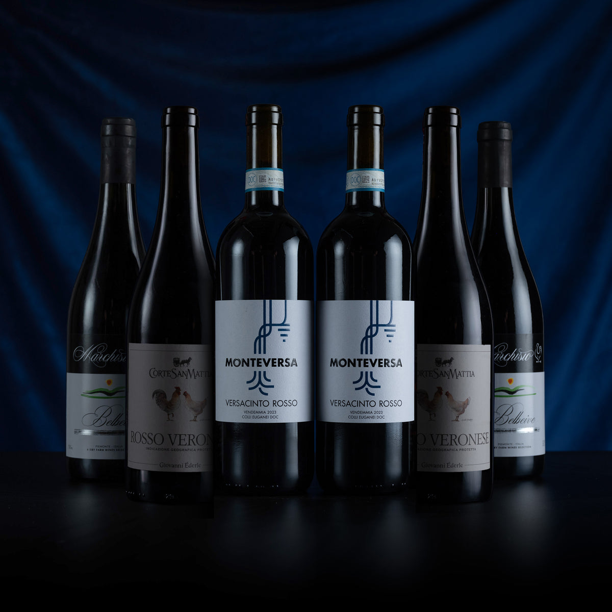 Six bottles of Monteversa red wine arranged in a row against a dark blue draped fabric background. The center bottles feature white labels with blue text and graphics, while the surrounding bottles have black labels.