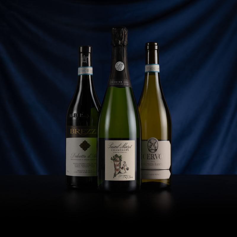 Three wine bottles arranged side by side on a dark surface against a draped navy blue background. The center bottle is a green sparkling wine bottle, flanked by a dark red bottle on the left and a pale yellow wine bottle on the right.