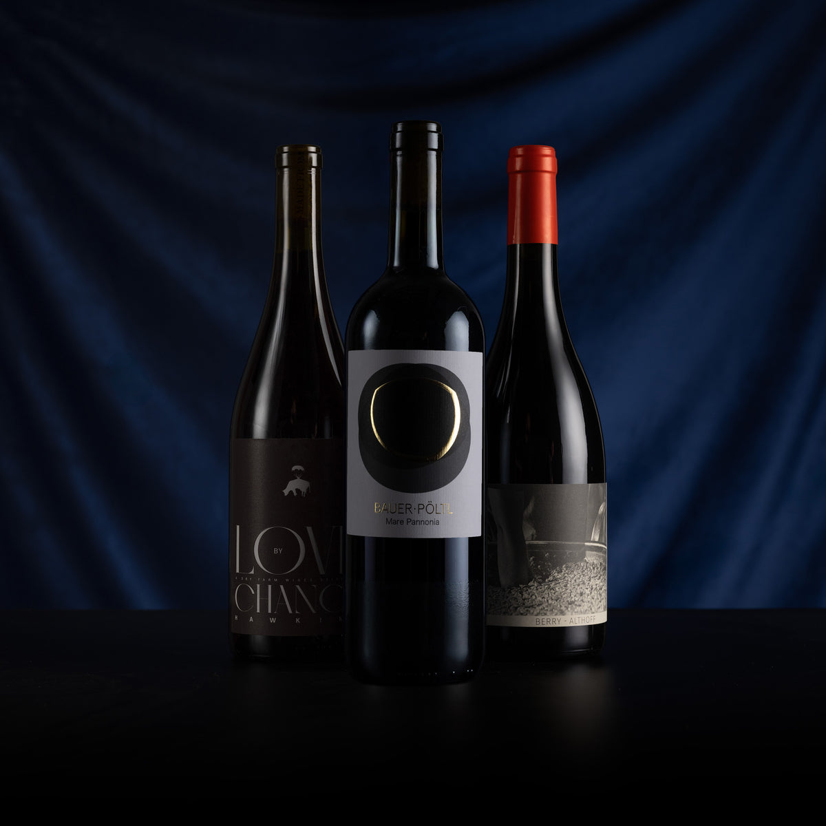 Three dark wine bottles standing on a black surface against a draped dark blue background. The center bottle has a white label with a large black oval and gold lettering, flanked by two dark bottles with black and red foil caps and dark labels.
